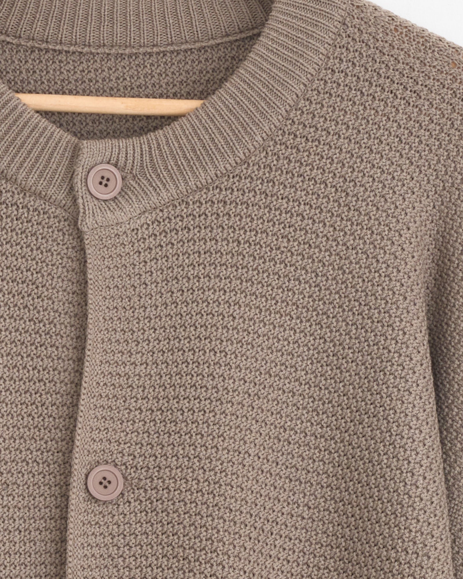 Close-up of the Rustic Knit Cardigan in Greige by Homme Plissé Issey Miyake on a hanger, featuring a ribbed collar, two brown buttons, and a waffle-knit texture. Need help with sizing or fit? Chat with us online!.