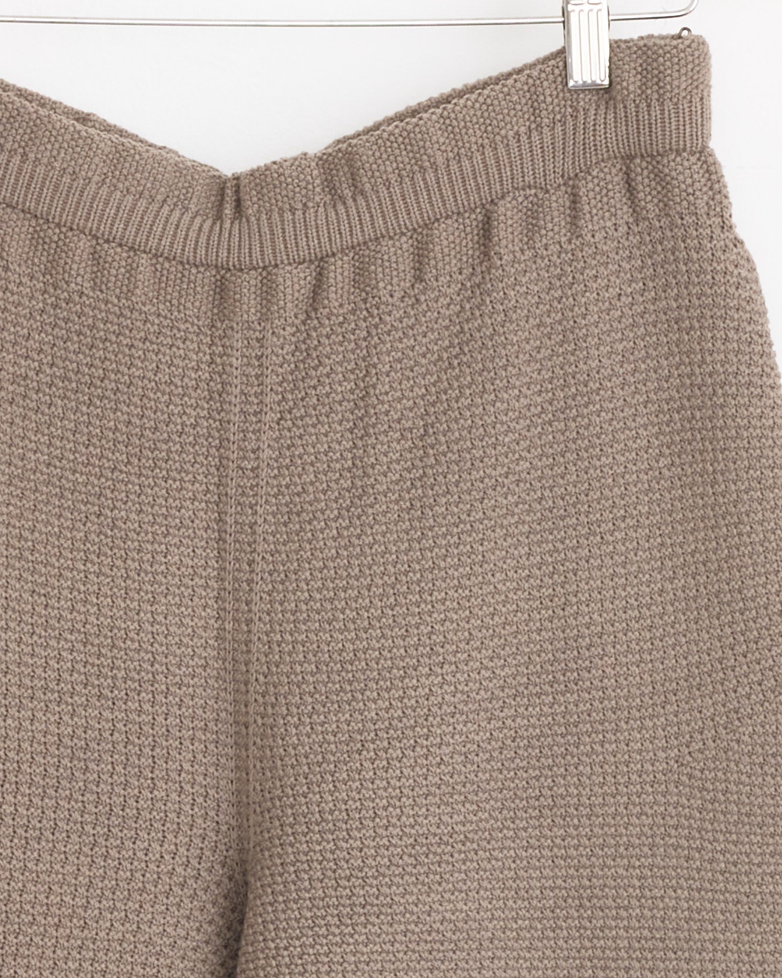 Close-up of the Rustic Knit Pant in Greige by Homme Plissé Issey Miyake, featuring an elastic waistband and soft, cozy fabric with a subtle waffle-textured knit, shown hanging against a white background.