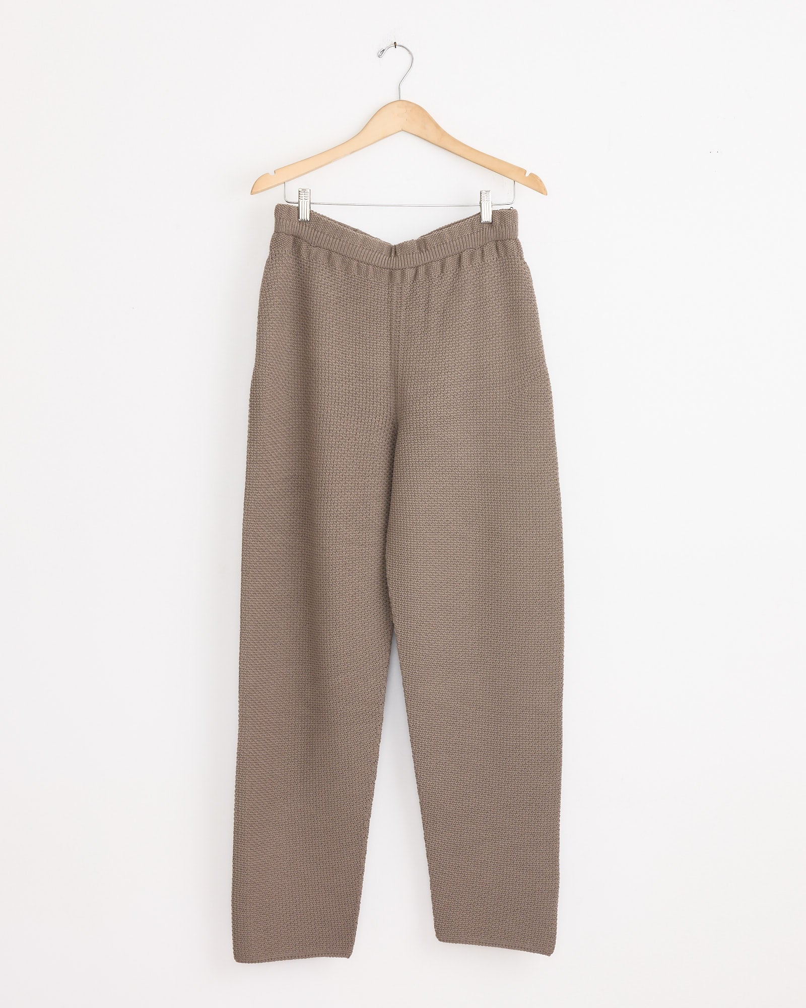 The Homme Plissé Issey Miyake Rustic Knit Pant in Greige, featuring a textured knit, elastic waistband, and relaxed straight-leg fit, hangs on a wooden hanger against a plain white wall.