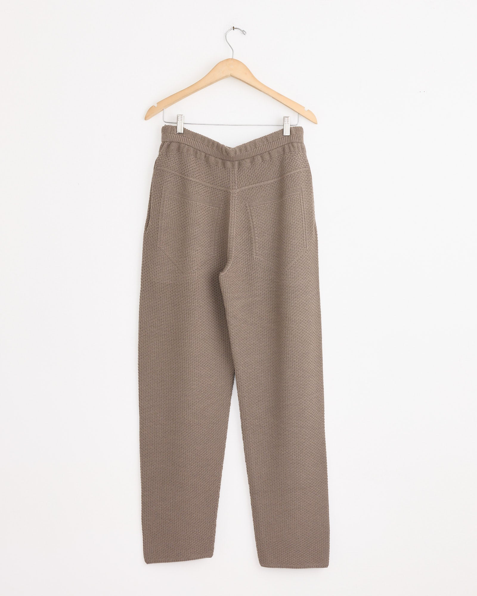 The Rustic Knit Pant in Greige by Homme Plissé Issey Miyake hangs on a wooden hanger against a white wall, featuring textured knit fabric, an elastic waistband, and side pockets.