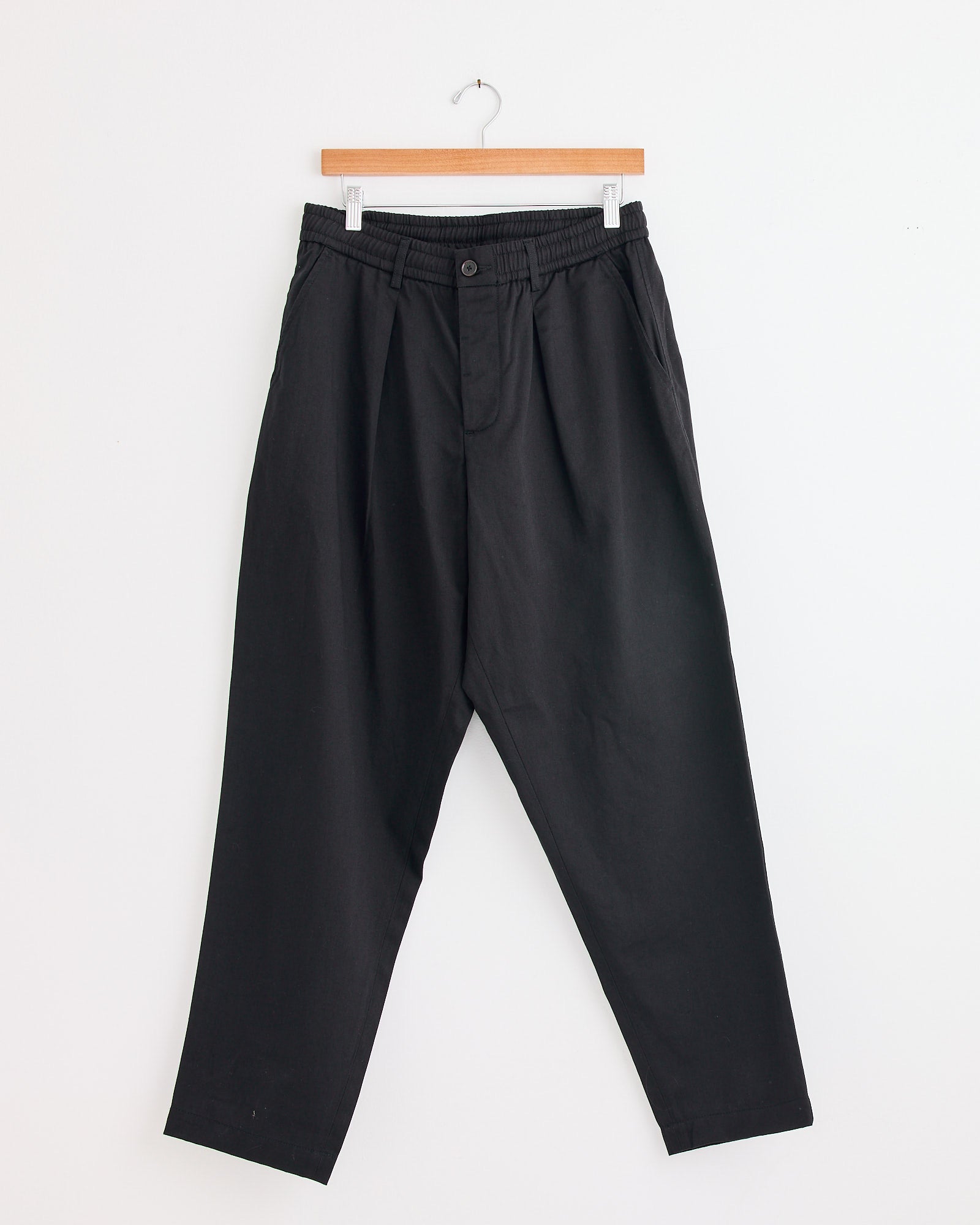 The Pleated Track Pant in Black by Universal Works hangs on a wooden hanger against a white wall. Features include an elastic waistband, side pockets, and a button closure. For fit or sizing questions, contact us via online chat.