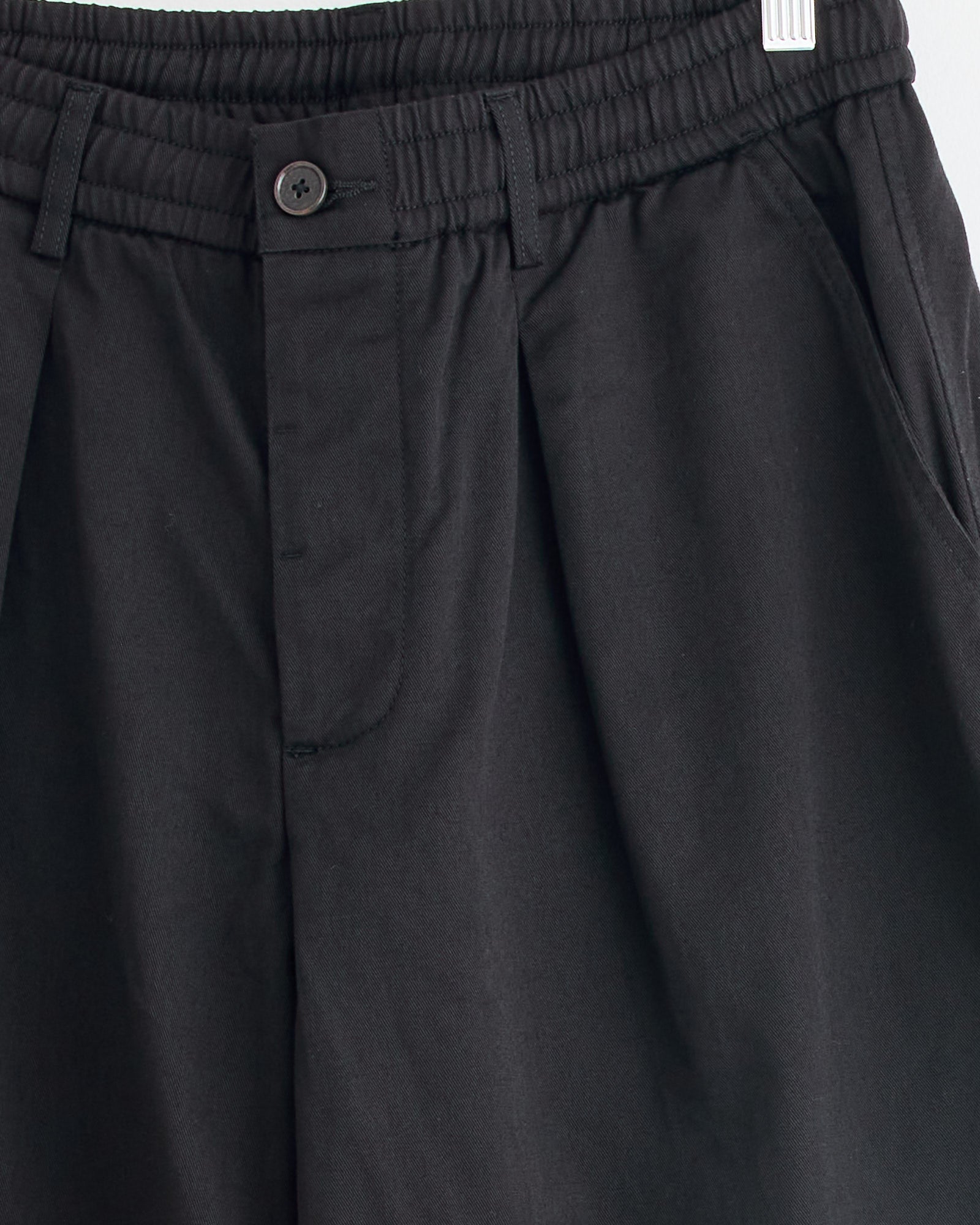 Close-up of Universal Works Pleated Track Pant in Black, featuring an elastic waistband, belt loops, button closure, and side pockets. Hanging on a white hanger against a light background. For fit questions, contact our online chat support.