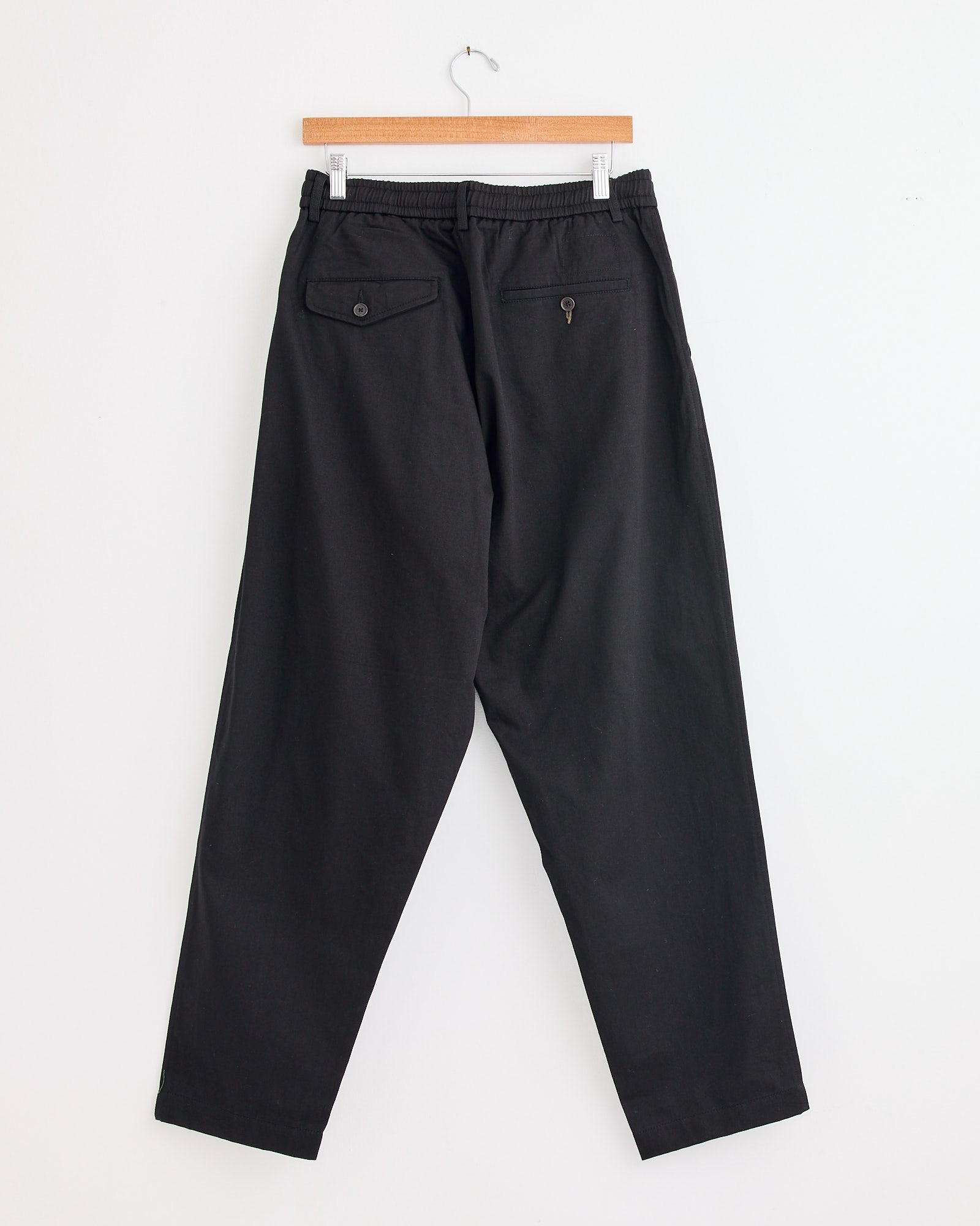The Pleated Track Pant in Black by Universal Works hangs on a wooden hanger against a white wall, shown from the back with two rear pockets—one with a button and one with a button loop—ideal for fit or sizing advice via online chat.