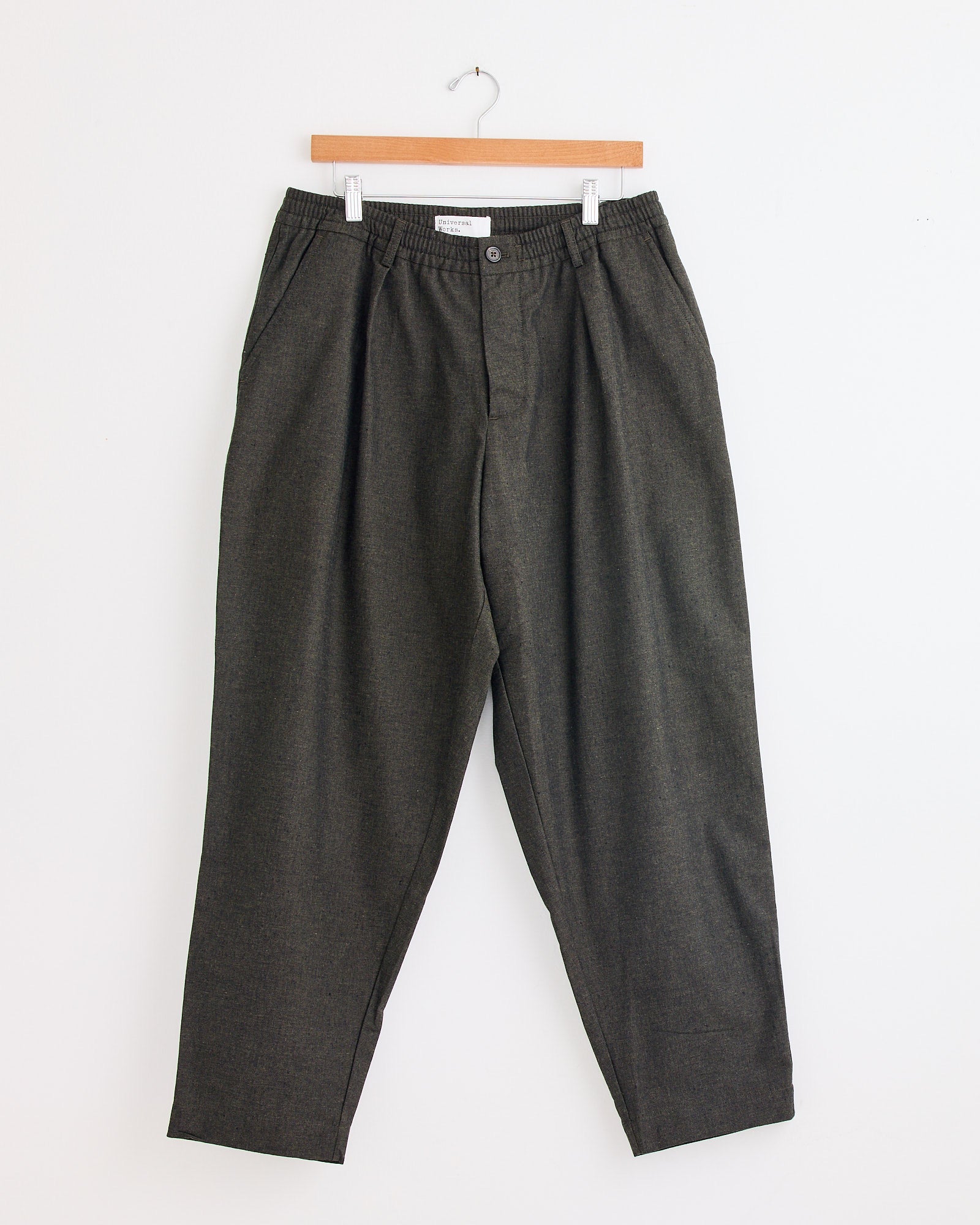 The Universal Works Pleated Track Pant in Olive features an elastic waistband, pleats, and side pockets, shown here on a wooden hanger against a white wall. For fit or sizing questions, please reach out via our online chat.