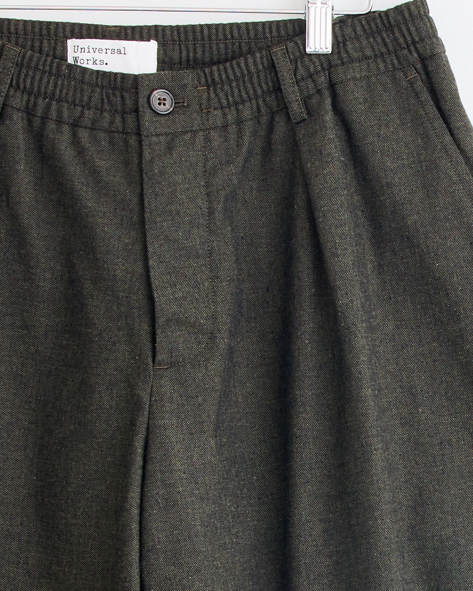 Close-up of Pleated Track Pant in Olive by Universal Works, featuring an elastic waistband, button closure, and relaxed fit, displayed on a metal hanger against a white background.