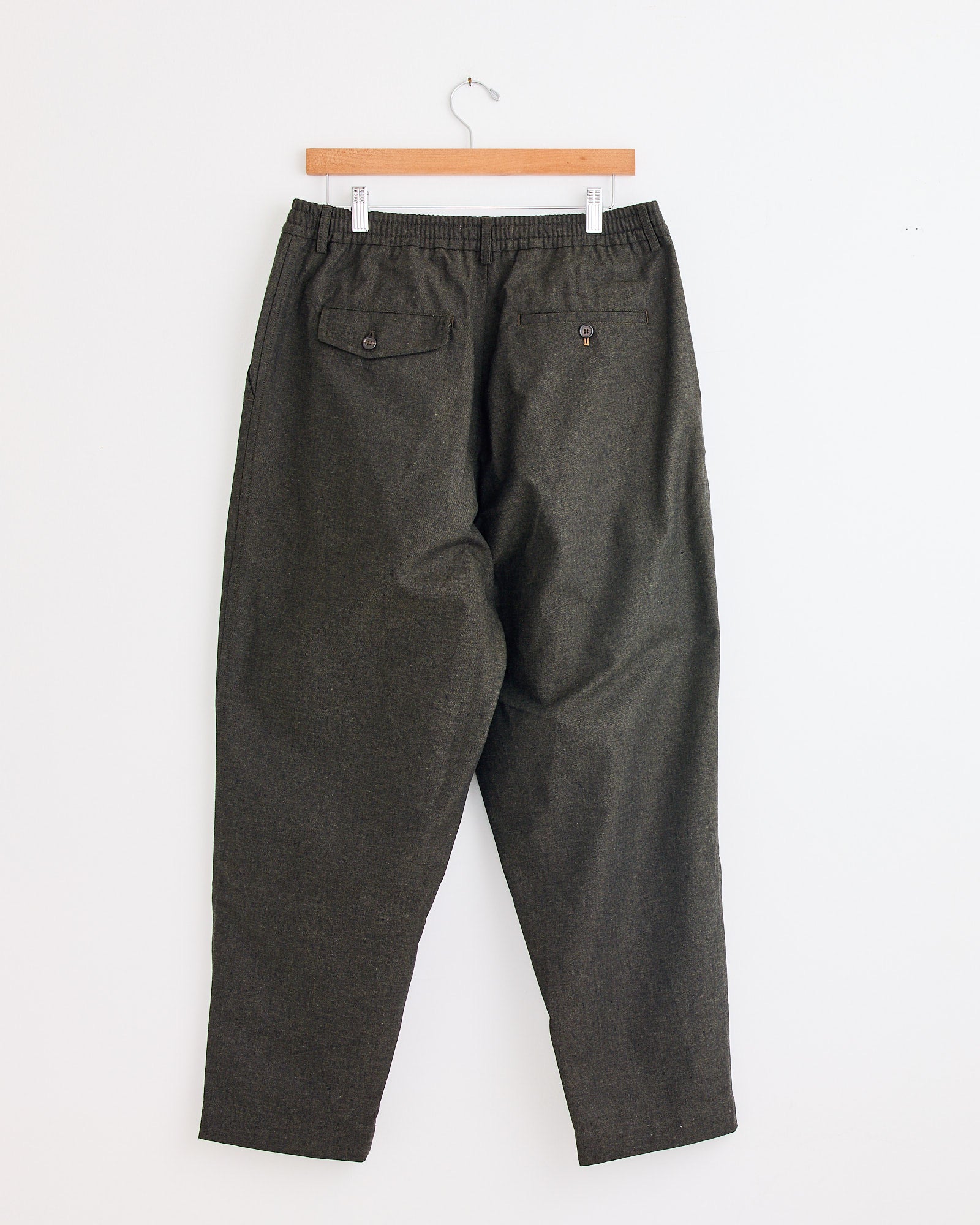 The Universal Works Pleated Track Pant in Olive, featuring a comfy elastic waistband and two buttoned back pockets, hangs on a wooden hanger against a white wall. Need help with sizing or fit? Chat with us online!.