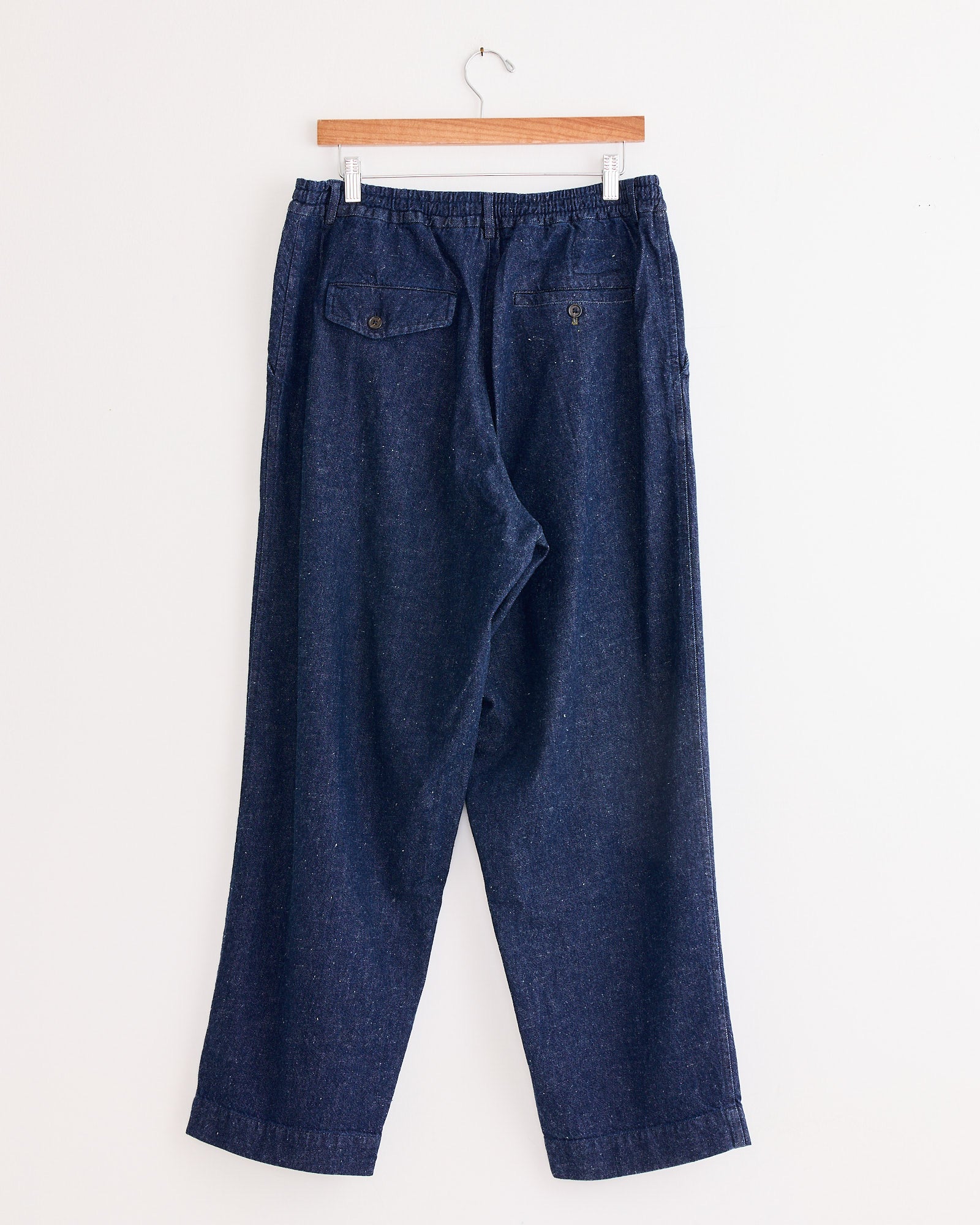 The Universal Works Oxford Pant in Indigo is a pair of loose-fitting, organic cotton trousers with an elastic waistband and two buttoned back pockets, shown hanging on a wooden hanger against a white background.