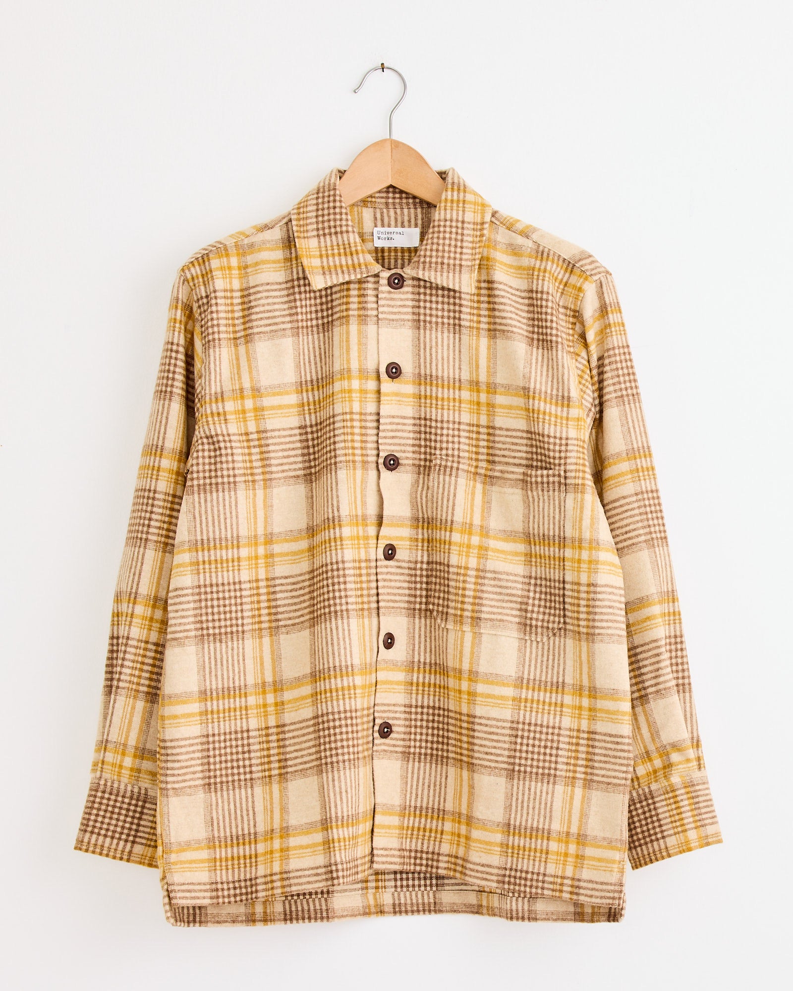 The Universal Works Fatigue Shirt in Gold features a beige plaid with brown and yellow checks, long sleeves, and brown buttons. Displayed on a wooden hanger against a white wall. For sizing help, contact us via online chat.