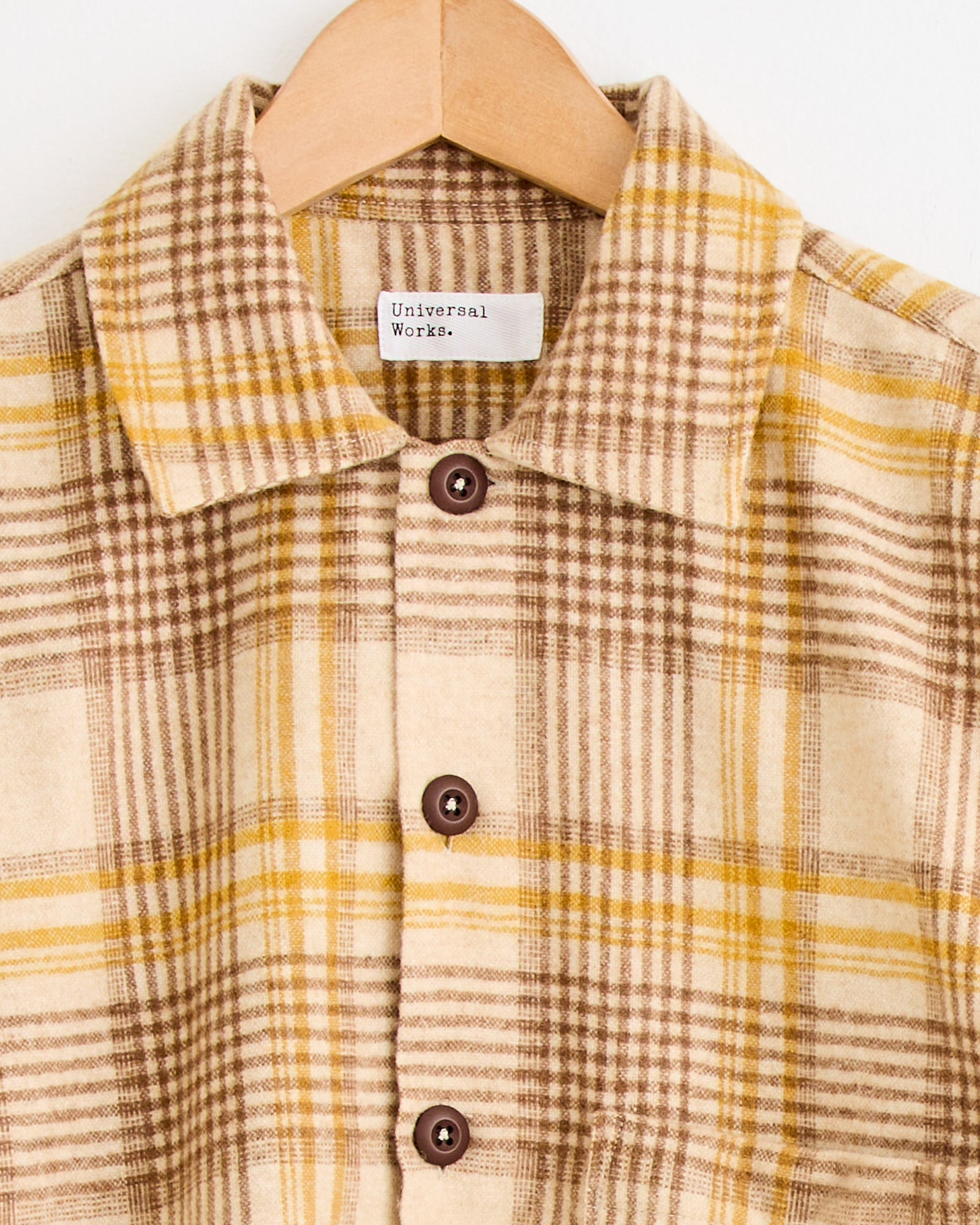 The Fatigue Shirt in Gold by Universal Works, featuring a beige and brown plaid with yellow accents, collar, and brown buttons, hangs on a wooden hanger. For the perfect fit, see our sizing guide or chat with us online.