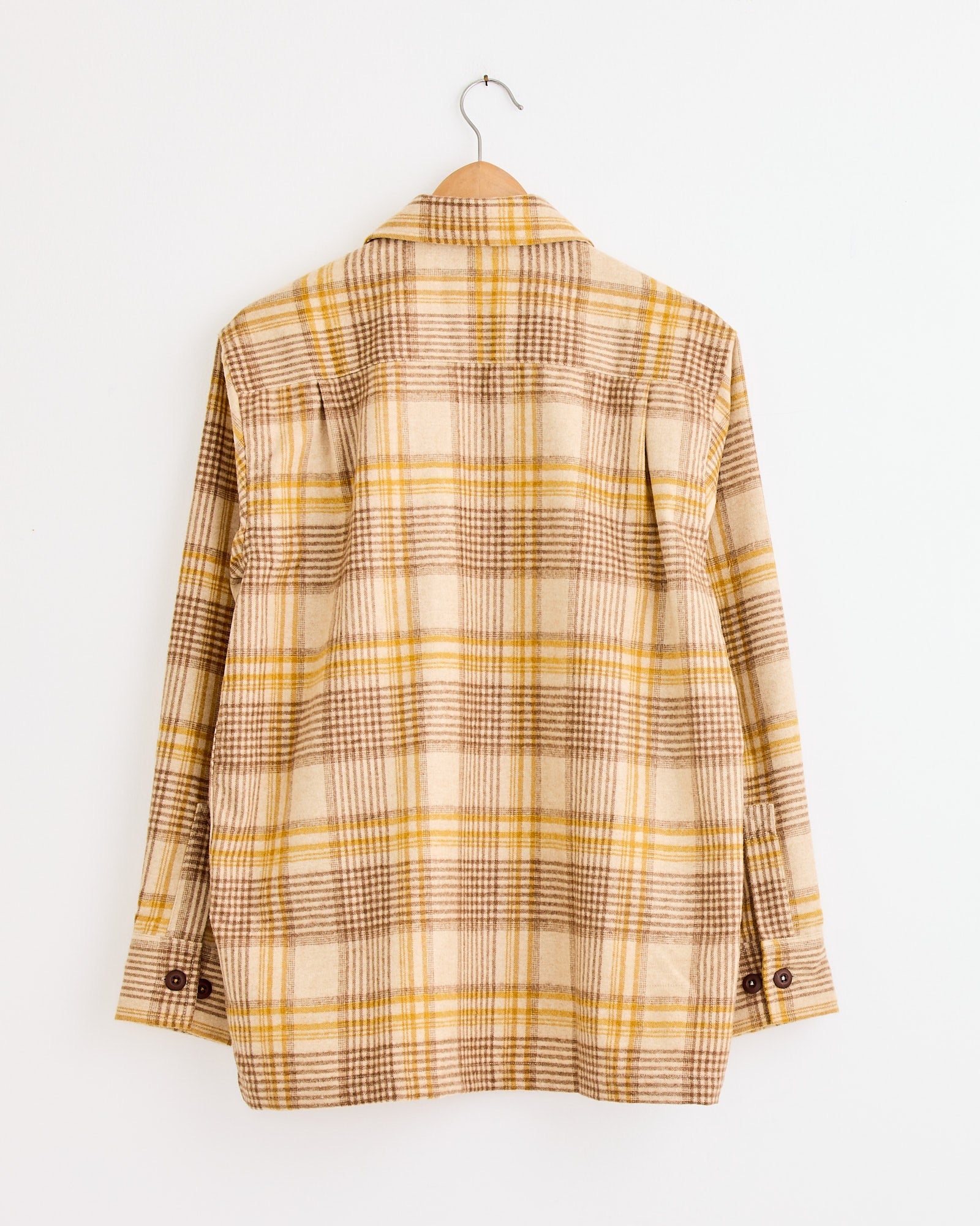 The Fatigue Shirt in Gold by Universal Works features a beige, brown, and yellow plaid flannel design, shown from the back on a wooden hanger against a white wall. For fit or sizing questions, please contact our online chat support.