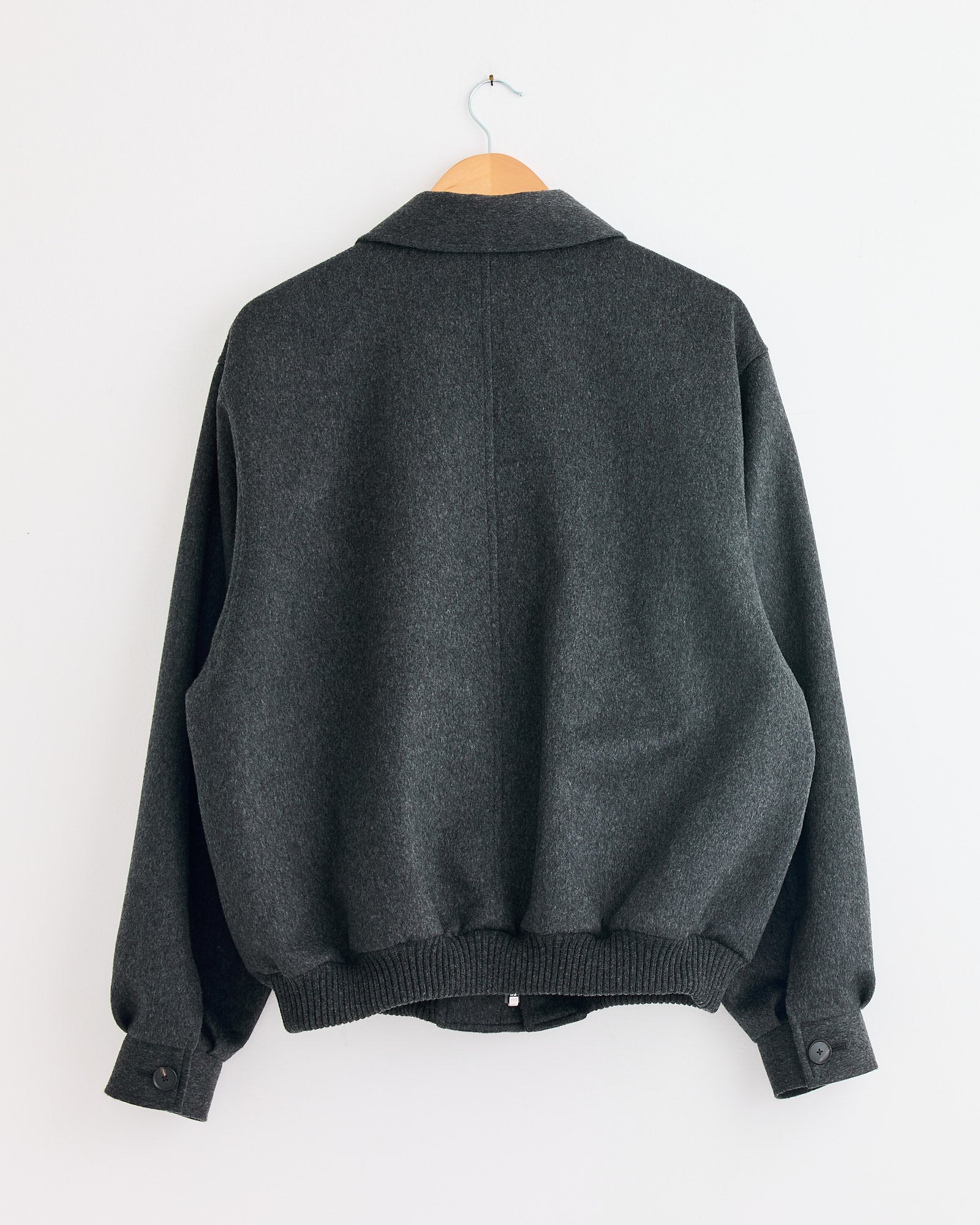 An Auralee Cashmere Moleskin Zip Blouson in Top Charcoal hangs on a wooden hanger against a white wall, shown from the back. Made in Japan, it features ribbed cuffs and hem with buttoned sleeves.