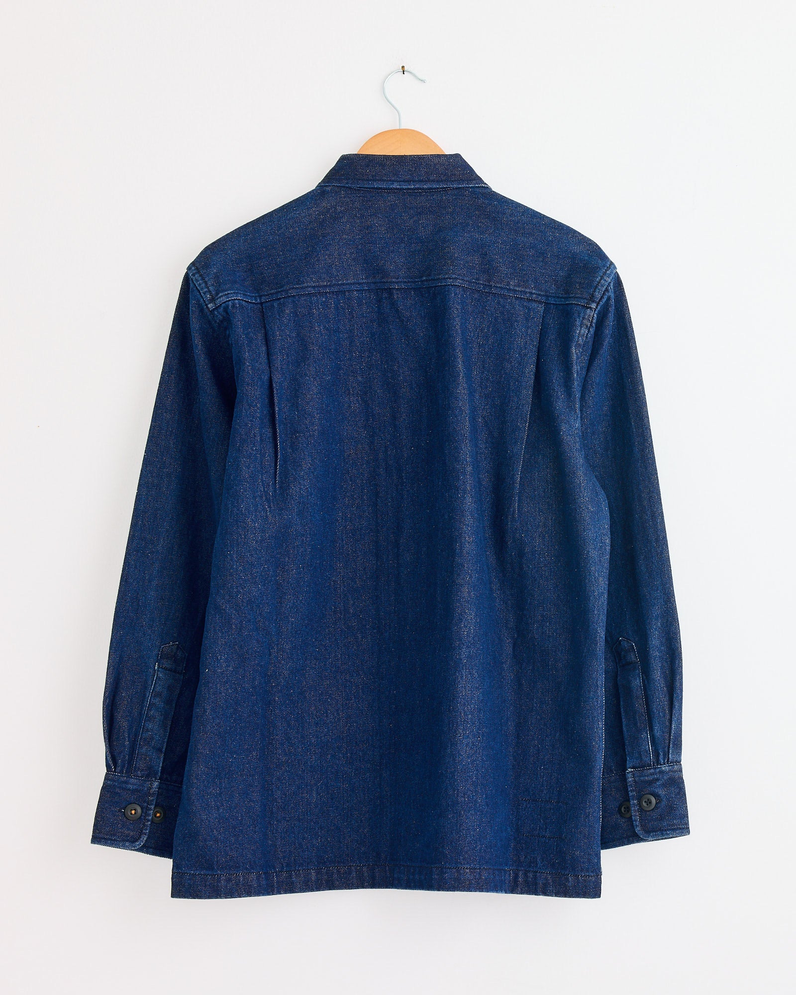 The Universal Works Fatigue Shirt in Indigo is displayed from the back on a hanger, highlighting its seams and buttoned cuffs—contact us via online chat for fit and sizing details.