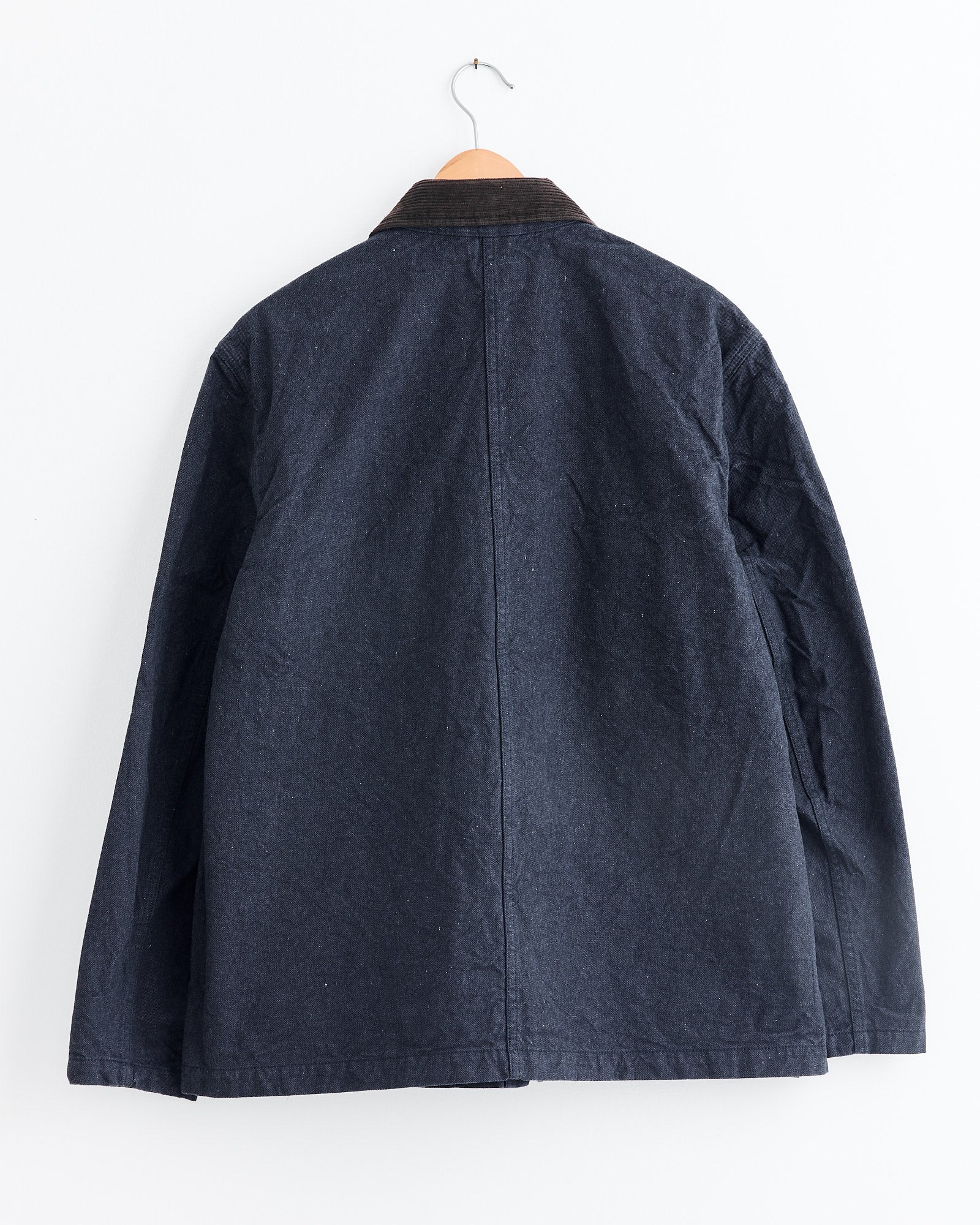 The Utility Jacket in Indigo by Universal Works features a brown corduroy collar and is shown from the back, hanging on a wooden hanger against a white wall. For sizing or fit questions, our online chat team is ready to help.