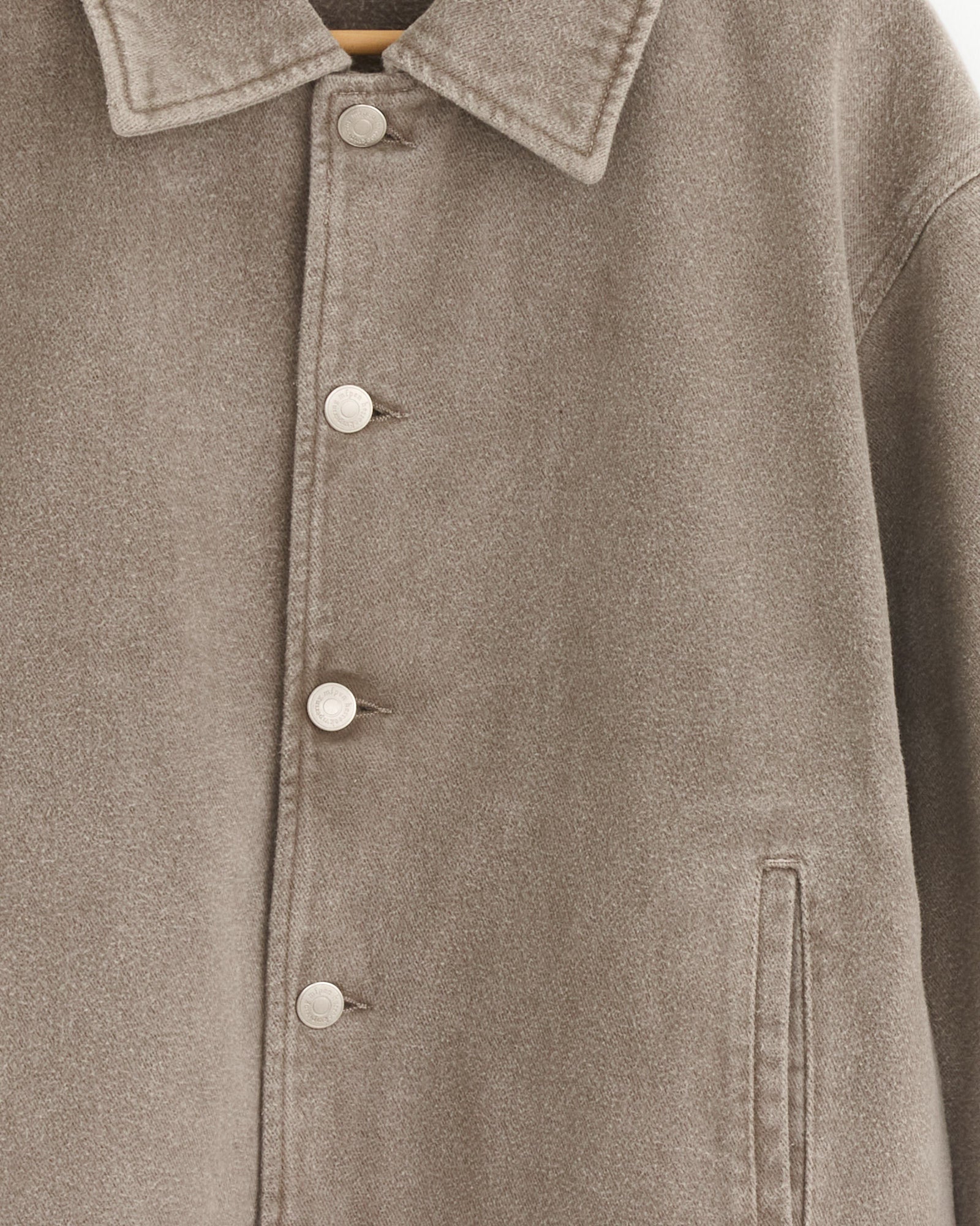 Close-up of the mfpen Painters Jacket Brushed in Mudstone—a beige wool jacket with a collar, silver buttons, and side pocket, hanging on a wooden hanger. The mineral dye washed finish lends subtle texture to this classic piece.