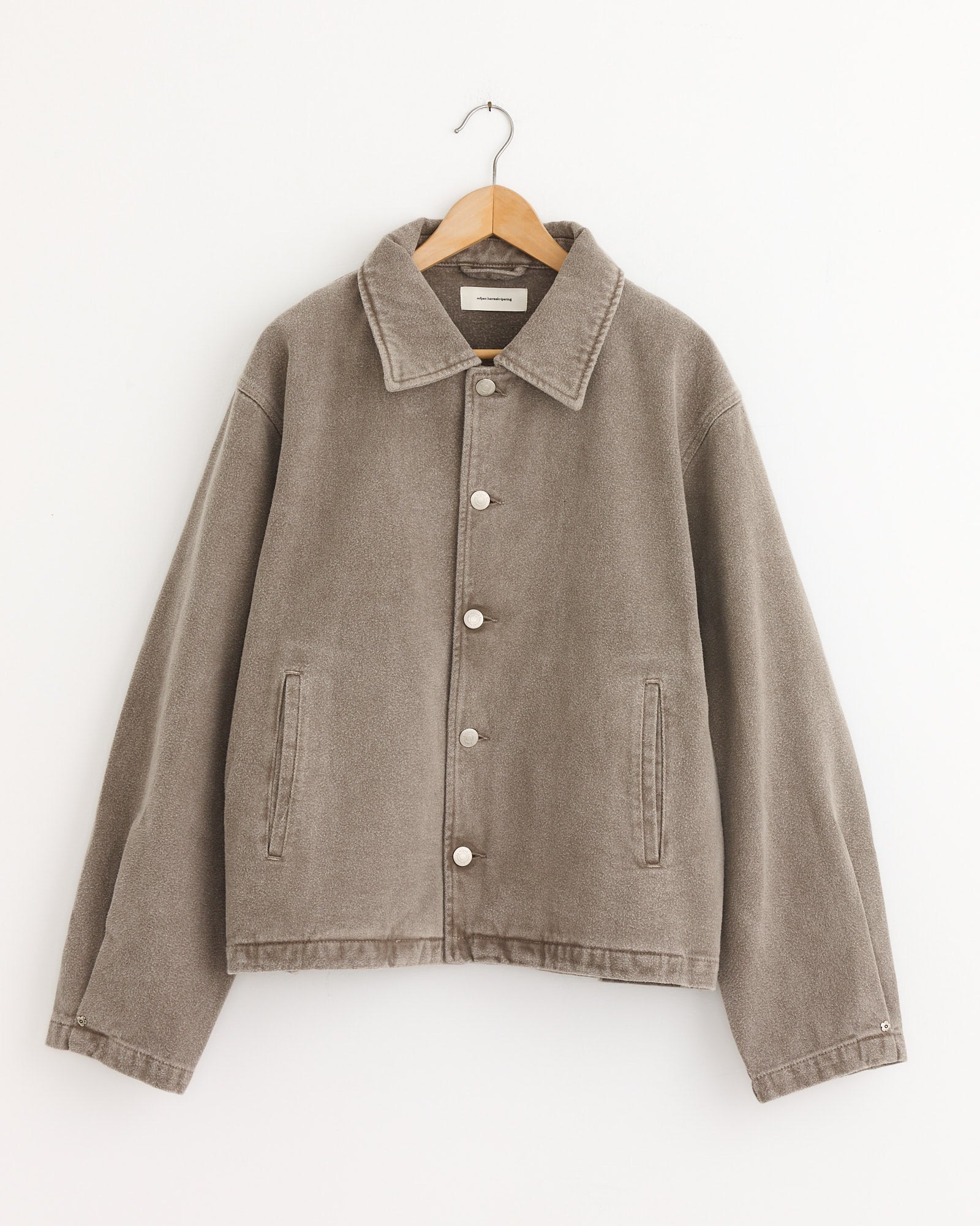 The mfpen Painters Jacket Brushed in Mudstone, made from organic cotton denim and mineral-dye washed, with a collar, button front, long sleeves, and two front pockets, hangs on a wooden hanger against a plain white backdrop.