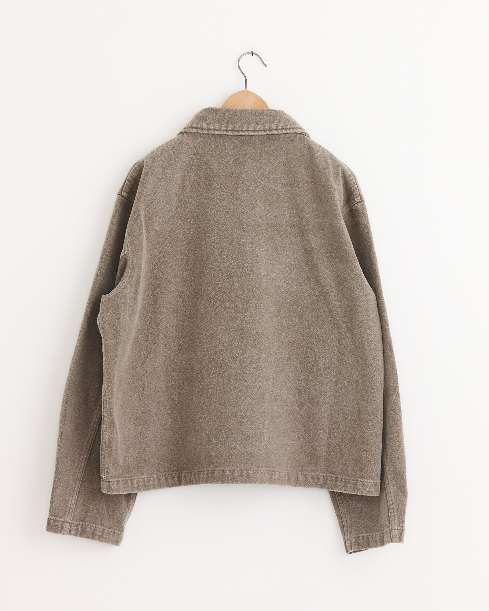 The mfpen Painters Jacket Brushed in Mudstone, a light brown piece with long sleeves and visible seams, is displayed on a wooden hanger against a plain white wall, shown from the back.