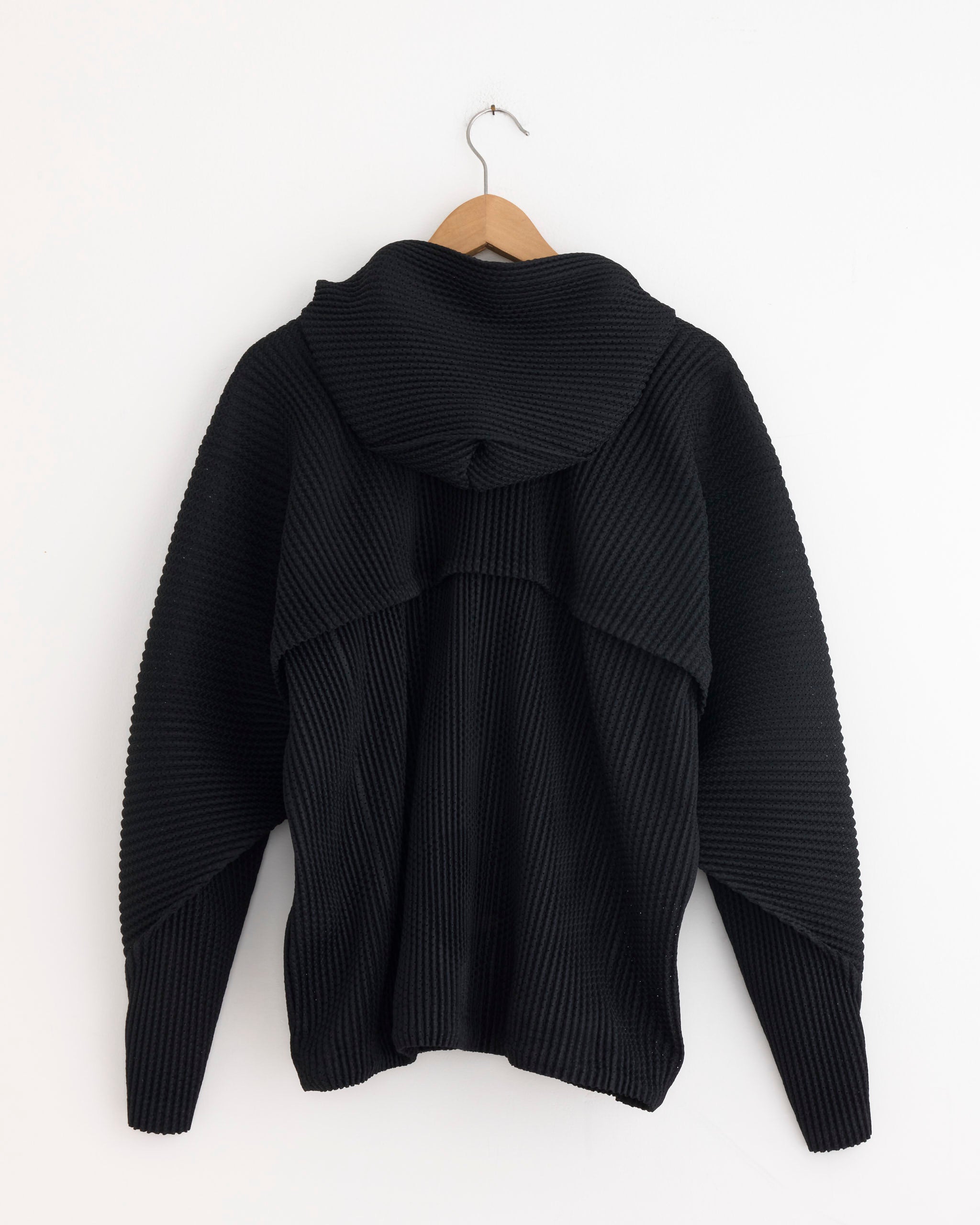 A black Homme Plissé Issey Miyake Wickerwork Mesh Jacket hangs on a wooden hanger against a white wall, displayed from the back.