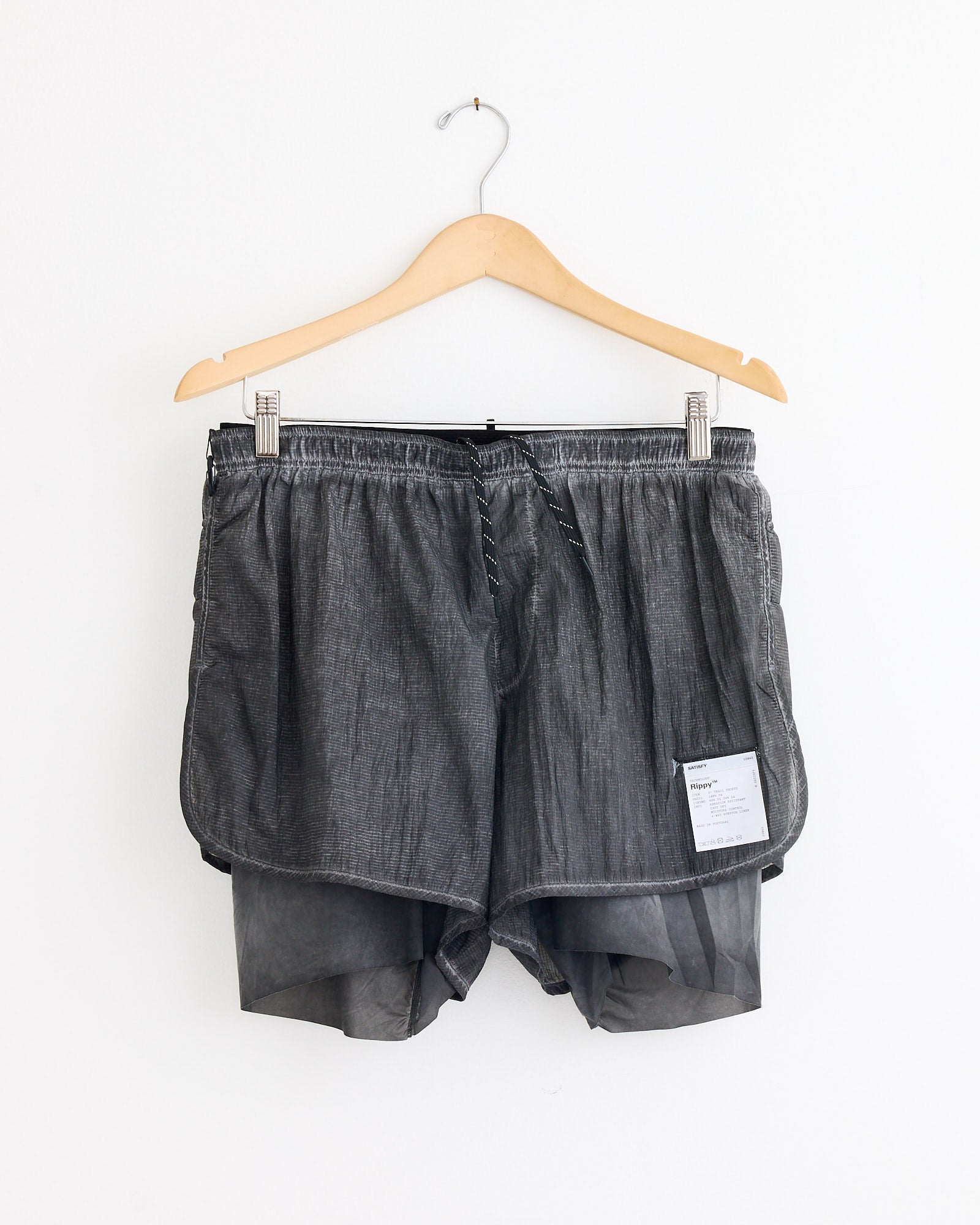 The Satisfy Running Rippy 3" Trail Short in Aged Black, featuring a sheer black inner lining and a small white label on the lower right, hangs on a wooden hanger against a white wall. For sizing questions, chat with us online.