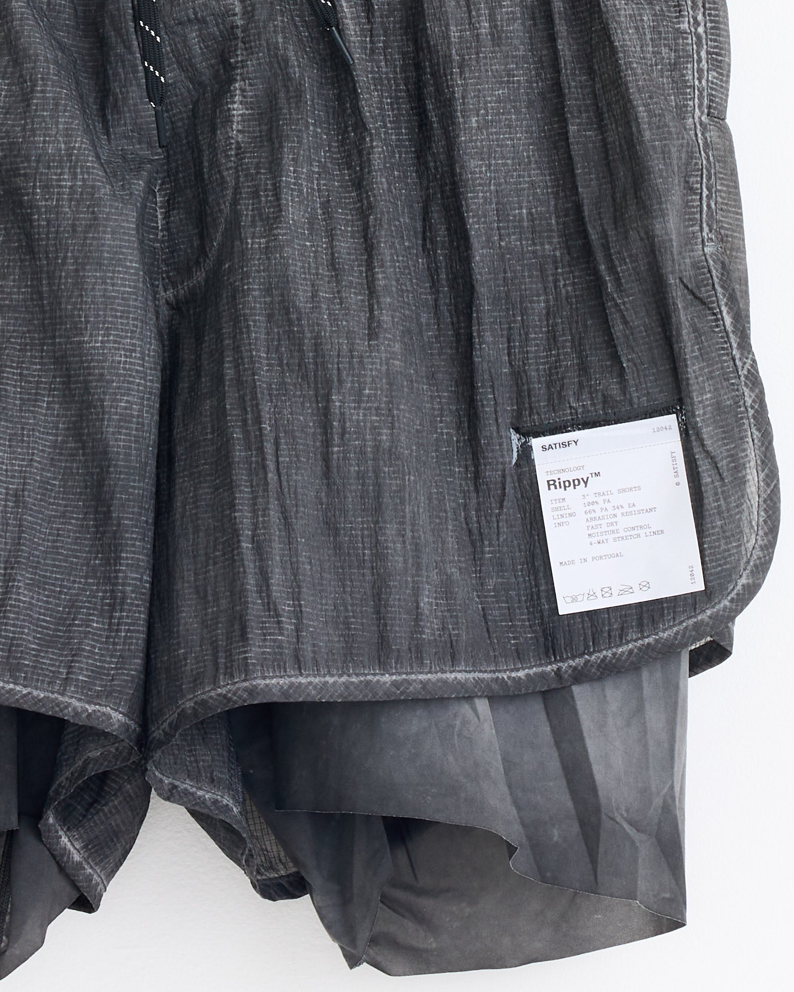 A close-up of Satisfy Running's Rippy 3" Trail Short in Aged Black, featuring a white label with text and barcode on the hem. The shorts are layered over a lighter inner lining for optimal fit and sizing.