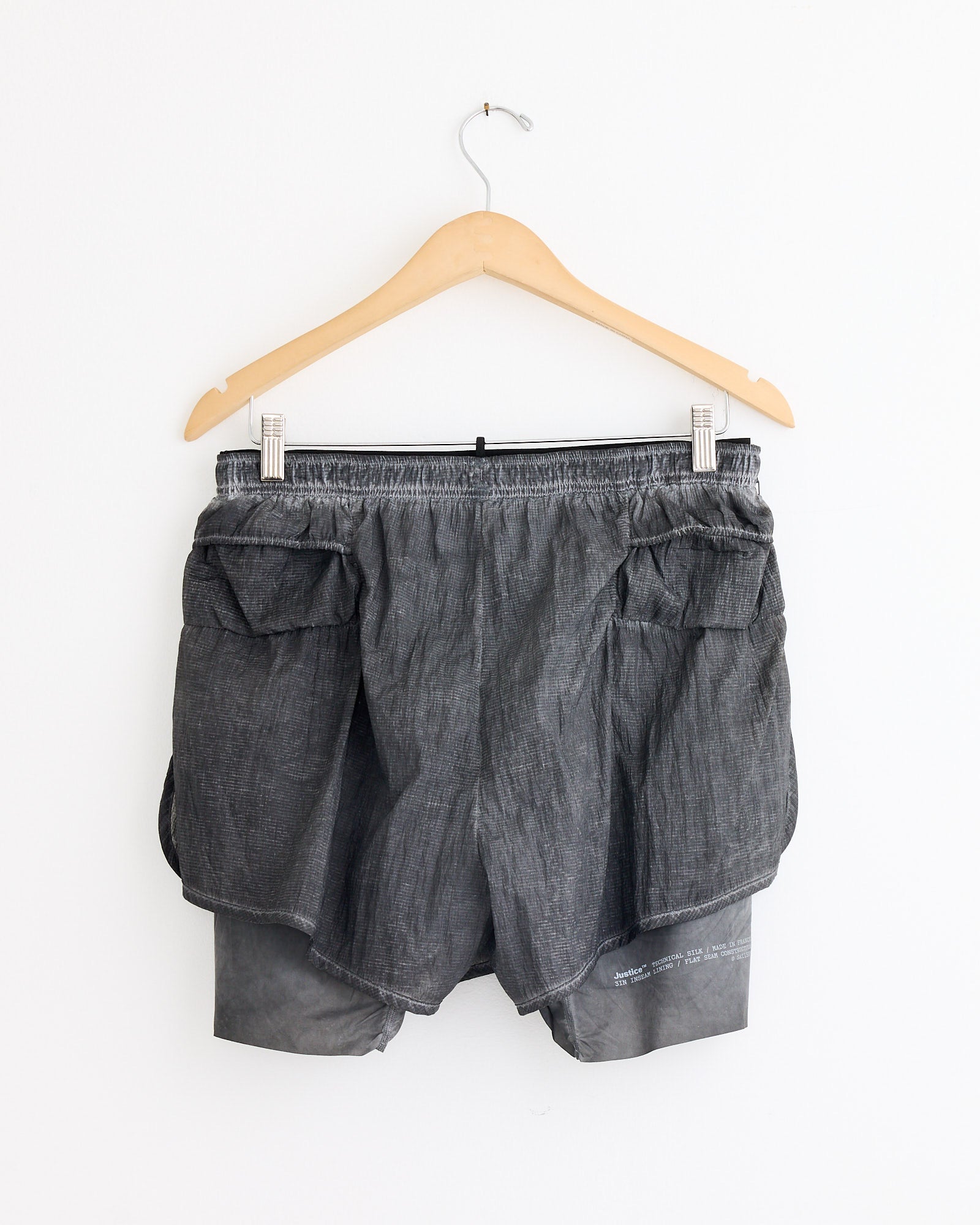 The Satisfy Running Rippy 3" Trail Short in Aged Black, shown on a wooden hanger against a white background, features multiple pockets—ideal for daily wear and available in versatile sizes for the perfect fit.