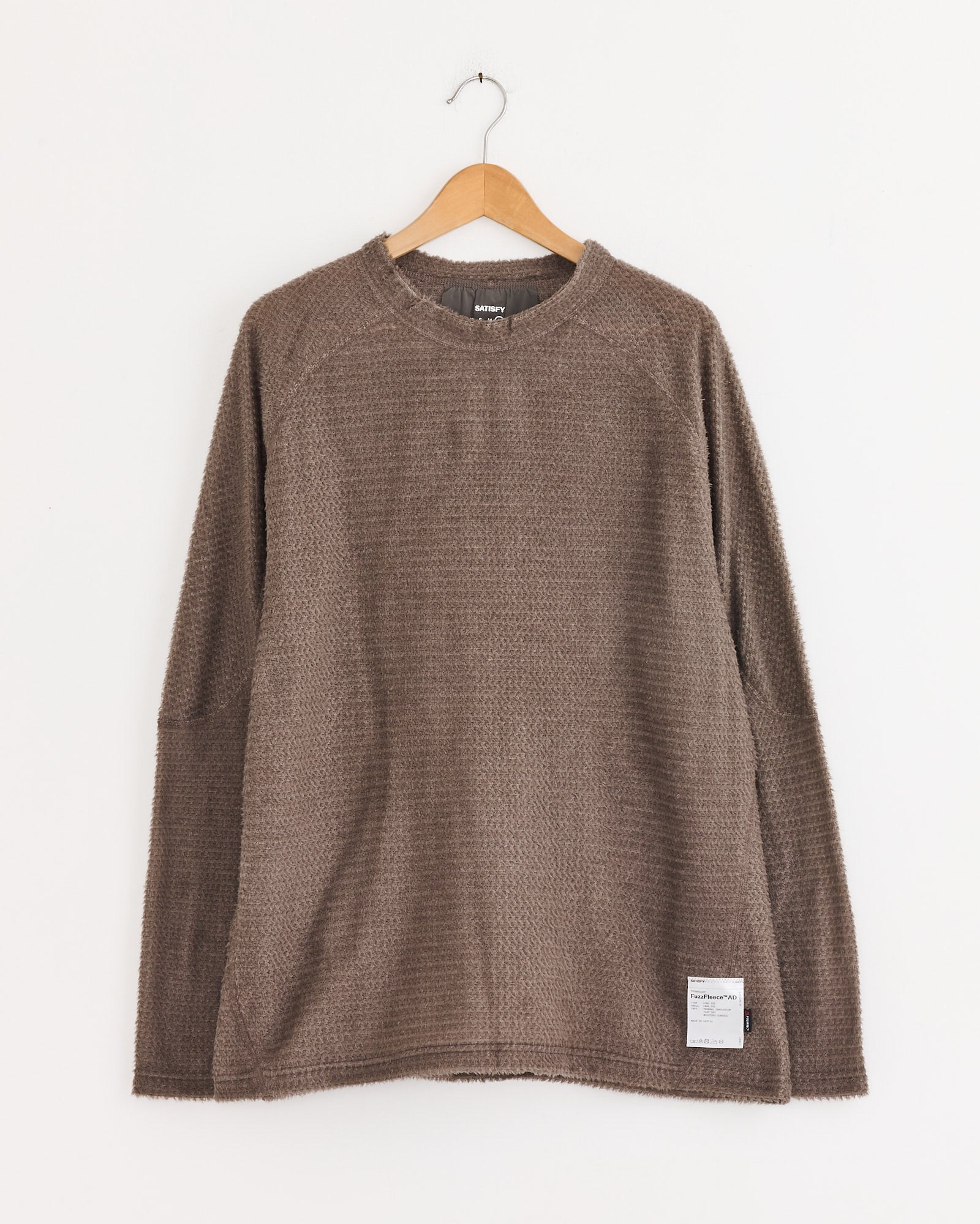 The Satisfy Running FuzzFleece AD Long Sleeve T-Shirt in Falcon, featuring textured fabric and moisture-wicking properties, hangs on a wooden hanger against a white wall. A small white label is sewn on the bottom left front.