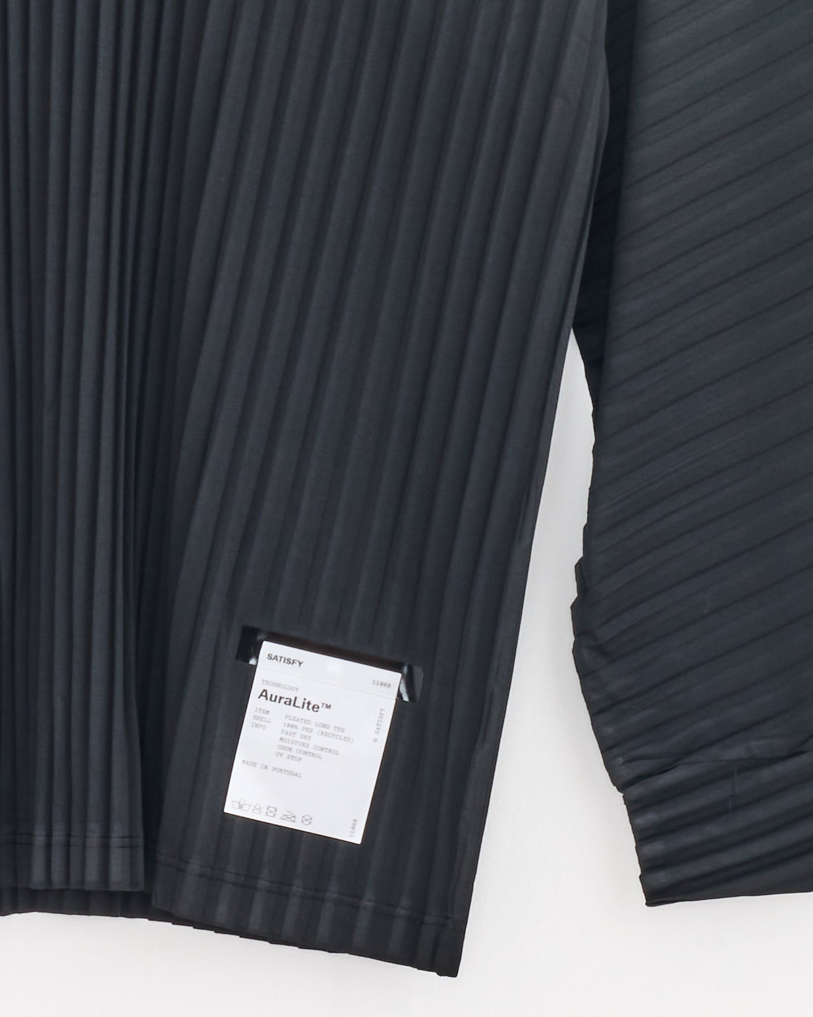 A close-up of the Satisfy Running AuraLite Pleated Long Sleeve T-Shirt in Black, made from recycled micro jersey, shows a visible white AuraLite™ tag sewn near the hem against a plain white background.