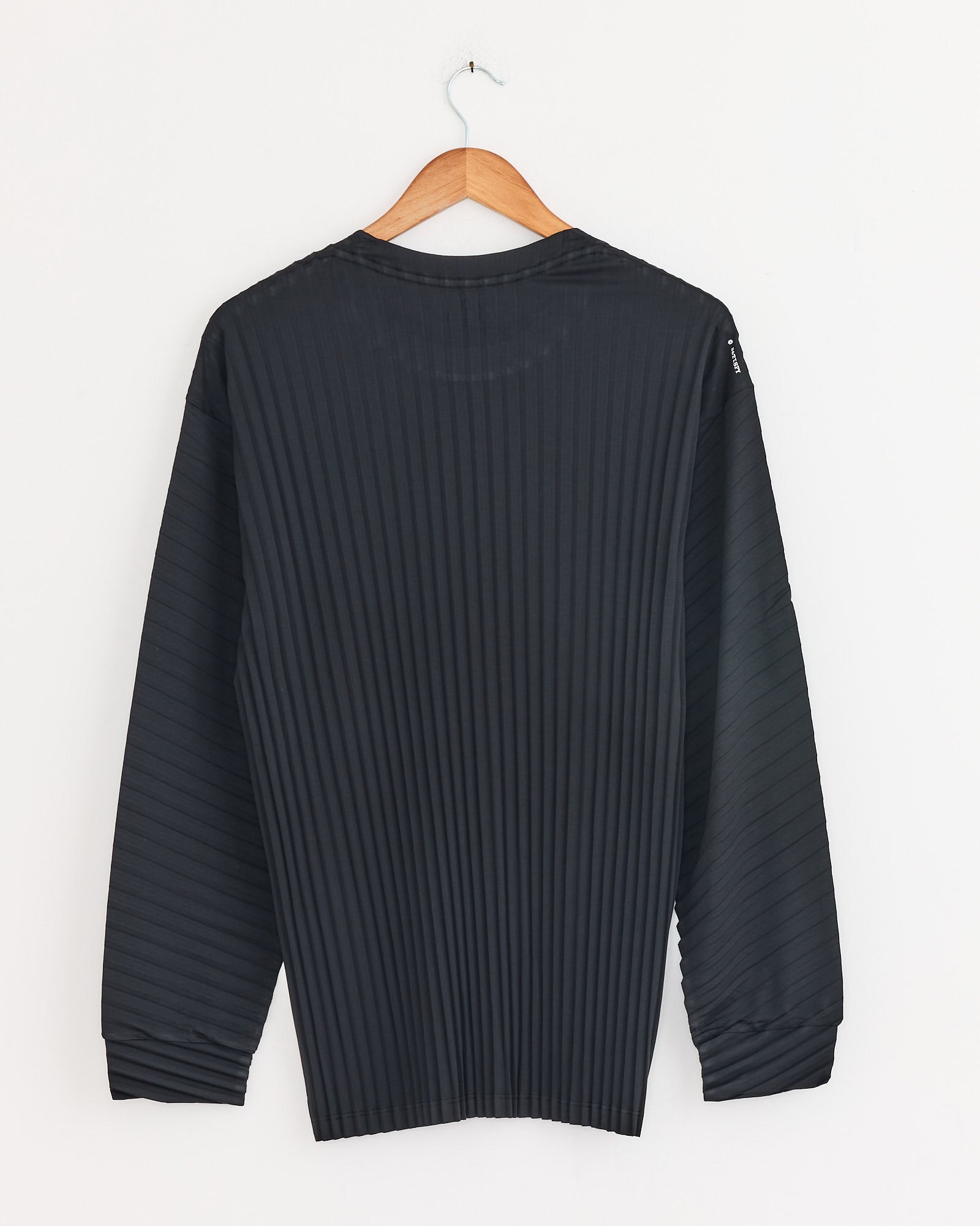 The Satisfy Running AuraLite Pleated Long Sleeve T-Shirt in Black, made from recycled micro jersey with moisture and odor control and a subtle vertical stripe pattern, hangs on a wooden hanger against a plain white wall, back facing forward.