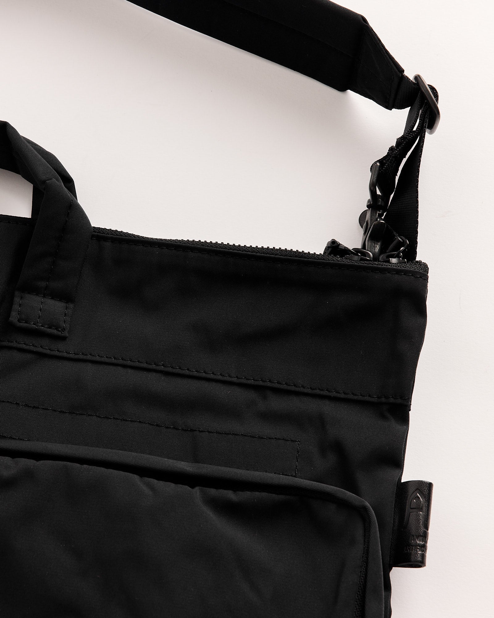 Close-up of the Amiacalva Gabardine Helmet Bag in Black, featuring a visible zipper, adjustable shoulder strap, and front pocket. Laid flat on a white surface, its sleek, minimalist design ensures an ideal fit and sizing.