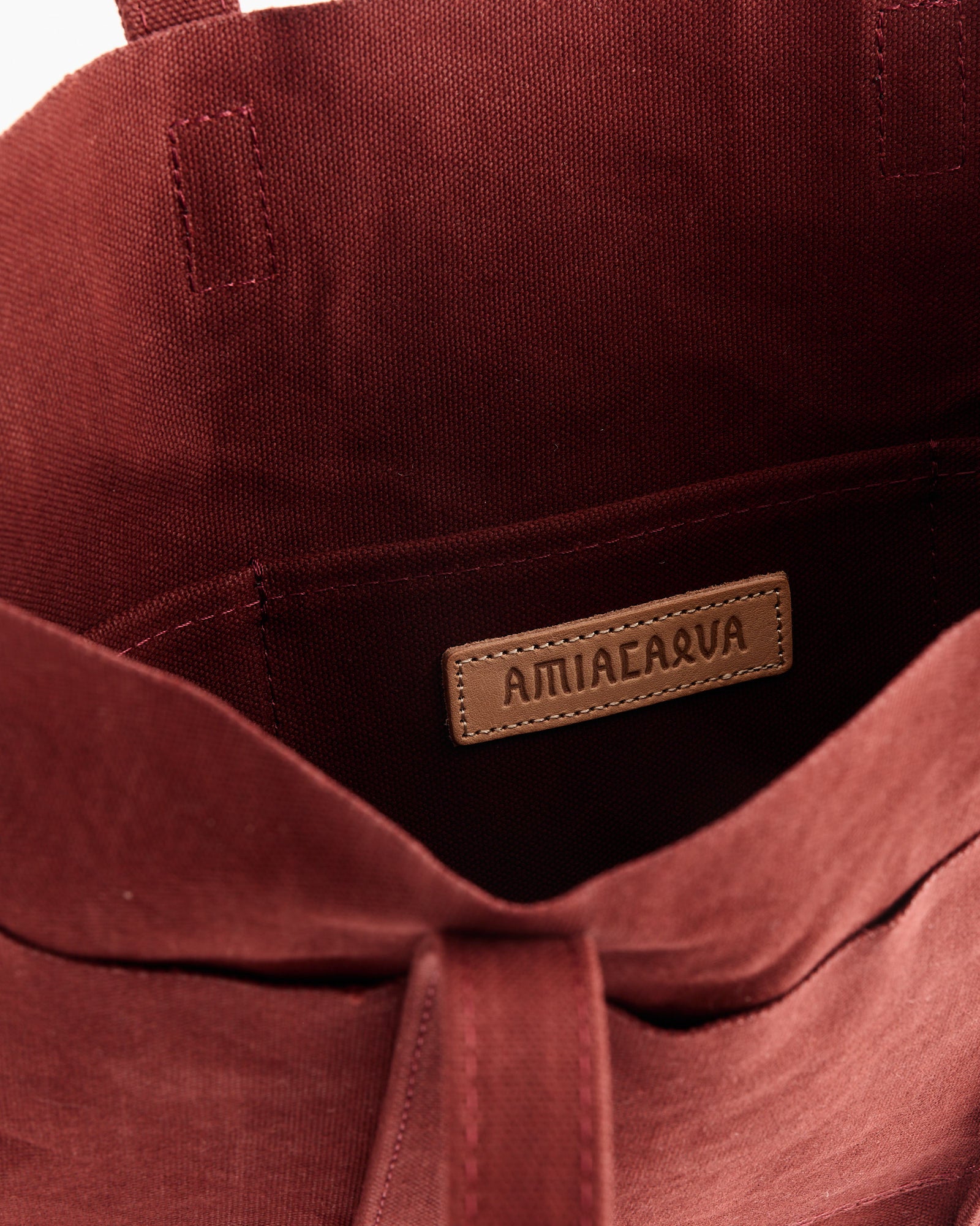 Close-up of the inside of the Washed Canvas 6 Pockets Small Tote in Burgundy by Amiacalva, featuring a stitched leather label with the brand name above an inner pocket.