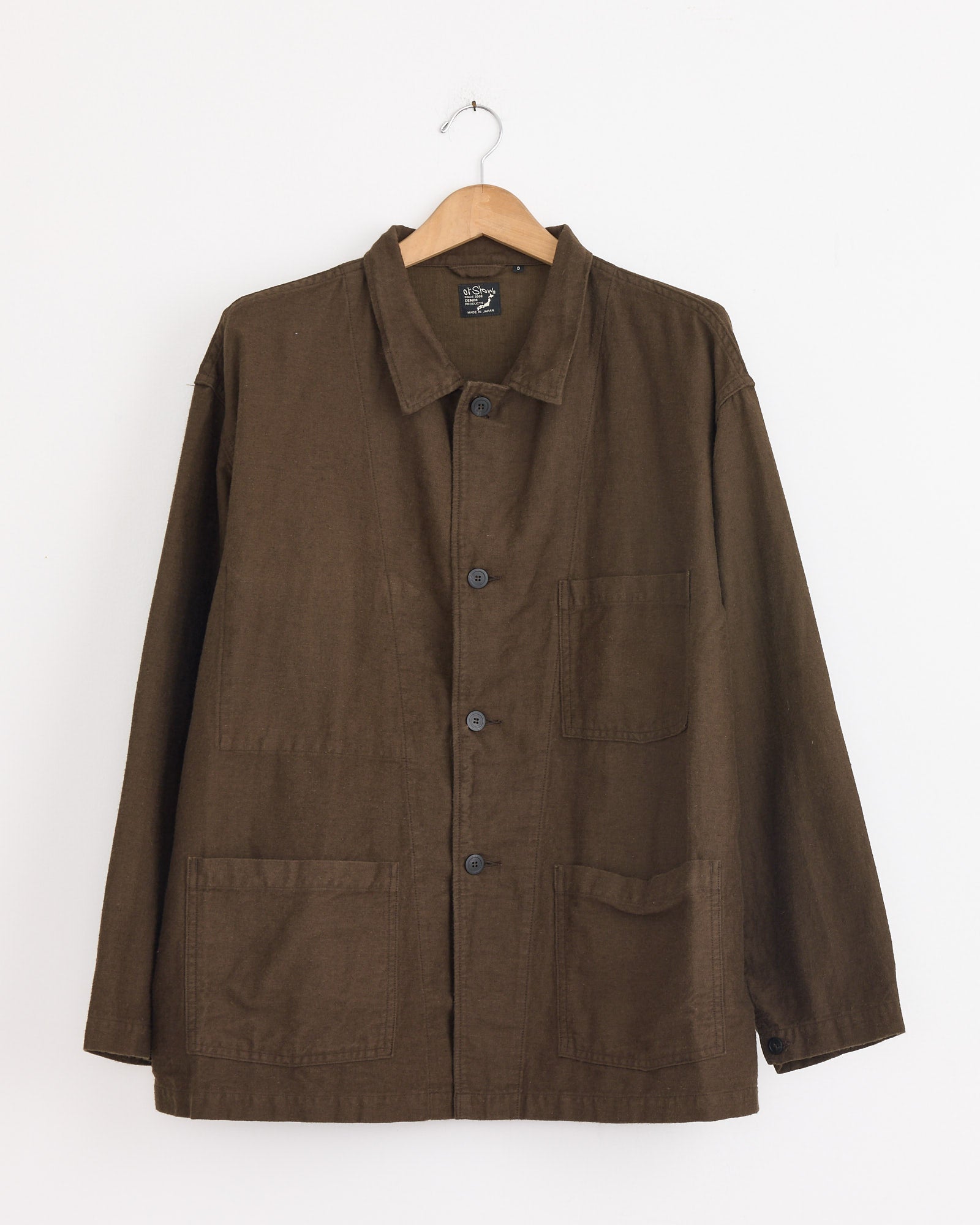 The orSlow Relax Fit Simple Work Jacket in Coffee Brown, featuring a collar and three large front pockets, crafted from cotton-linen moleskin, hangs on a wooden hanger against a plain white wall.