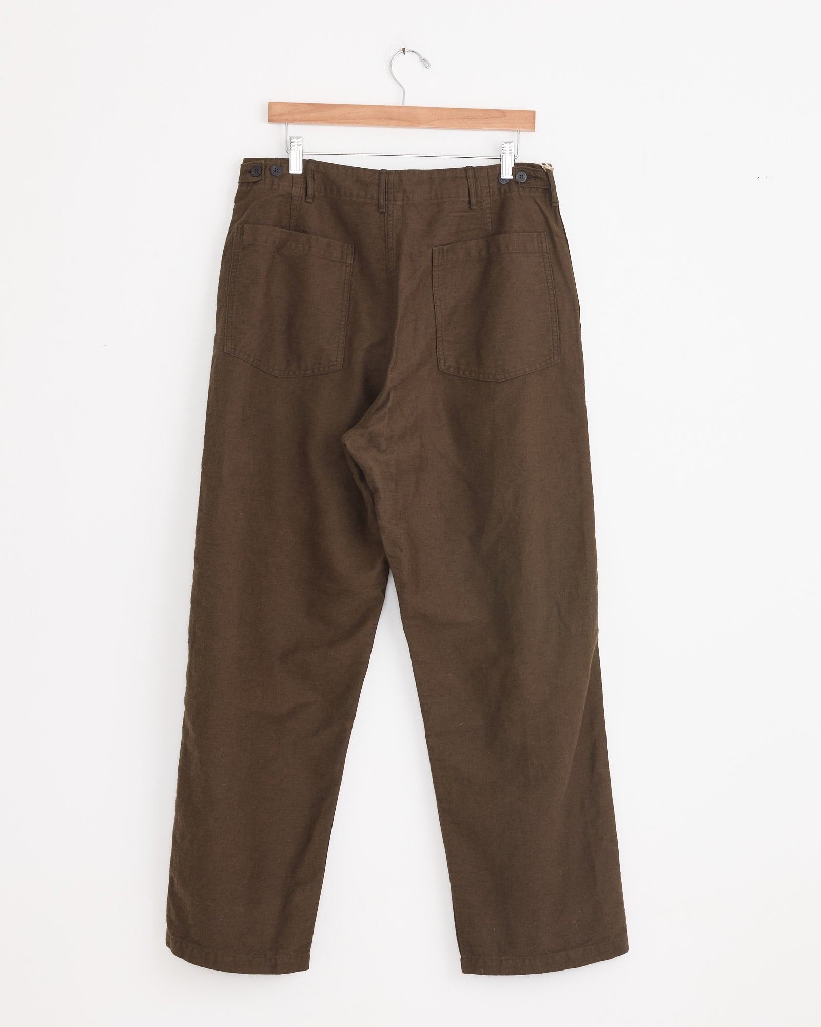 A pair of orSlow Relax Fit Fatigue Pant in Coffee Brown hangs on a wooden hanger against a white wall, featuring two back pockets.