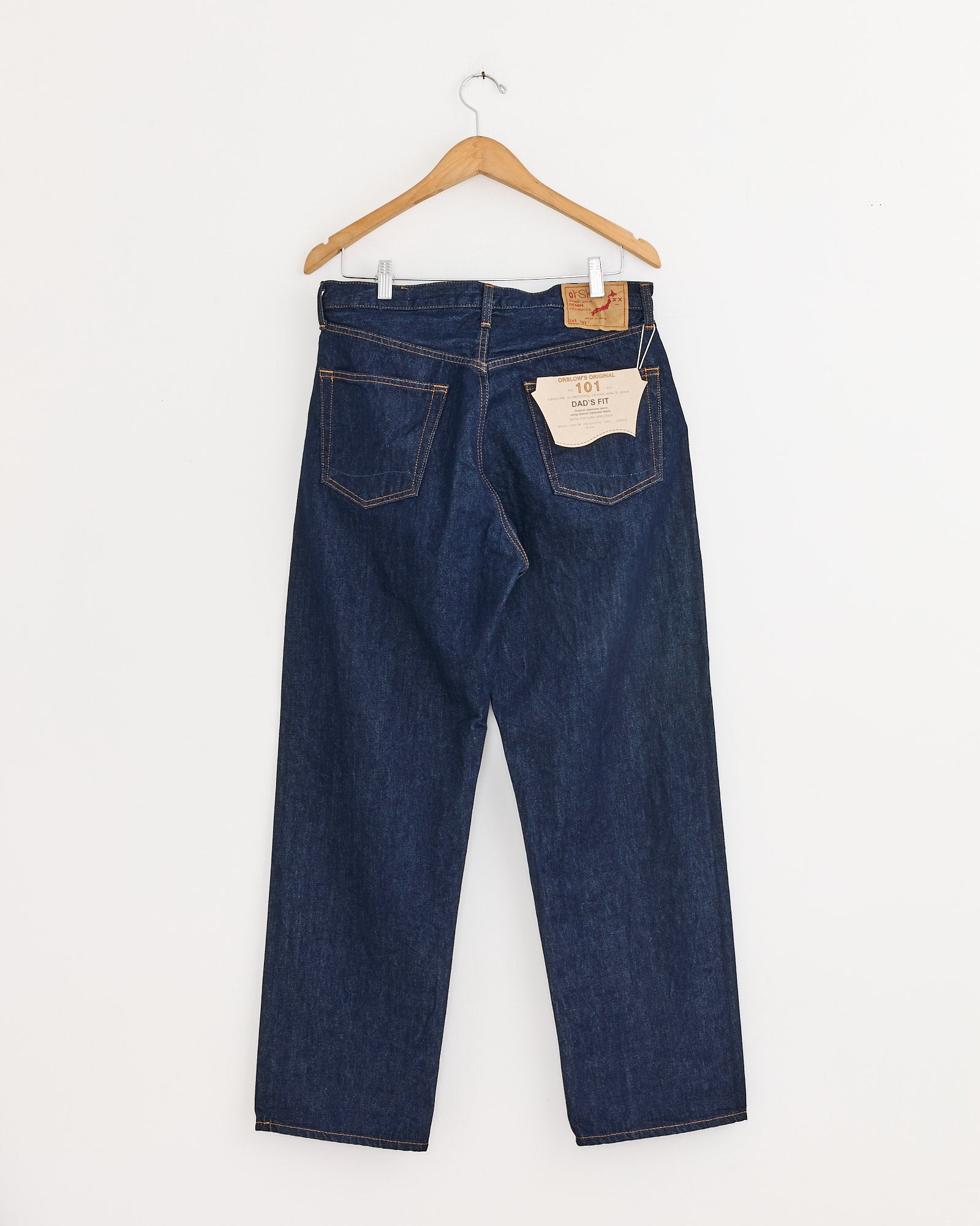A pair of dark blue Japanese denim jeans, the orSlow 101 Dad's Fit Denim Pant in One Wash, hangs on a wooden hanger against a white wall, featuring a brown patch and beige tag on the back waistband.