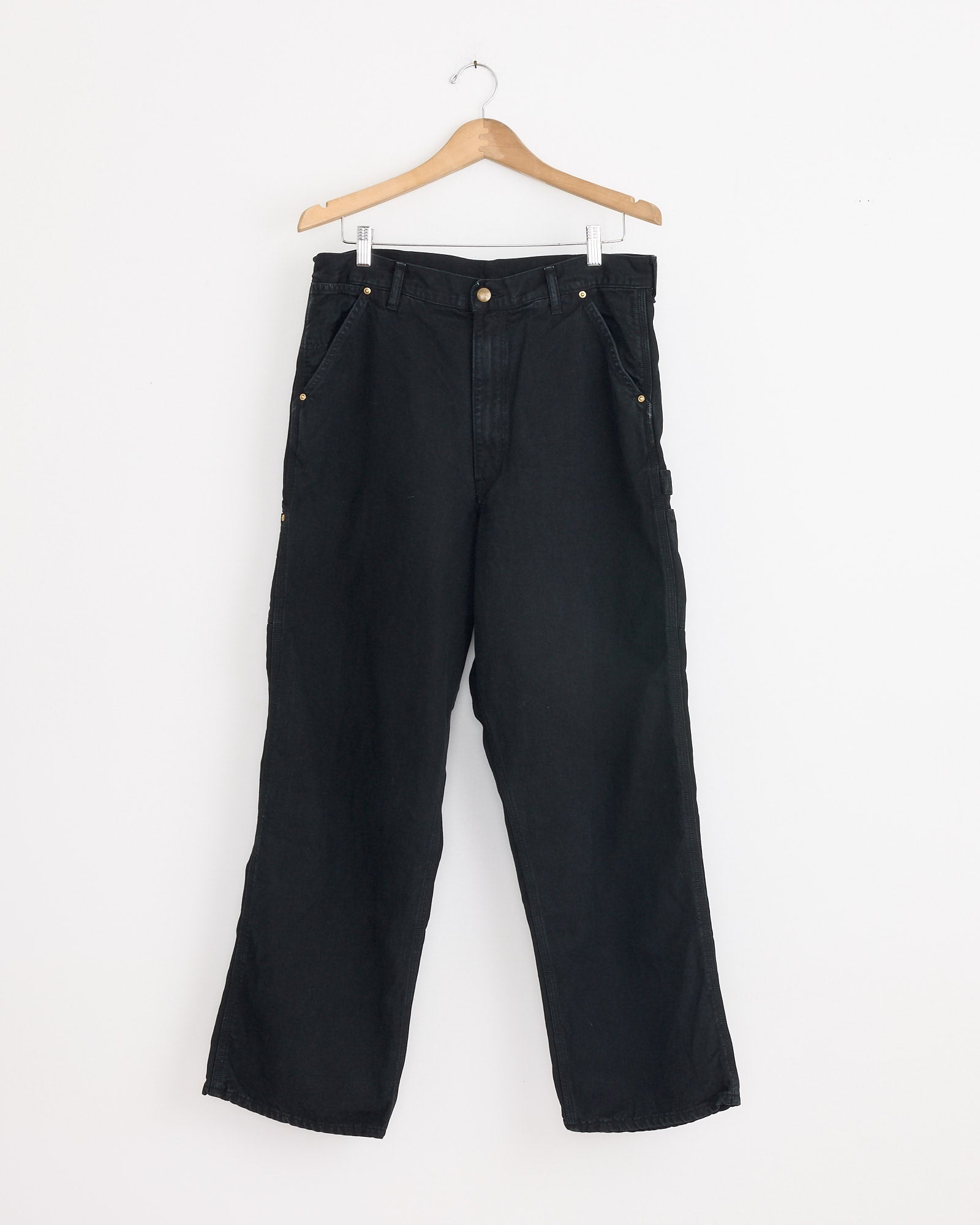 The orSlow Dad's Fit Oxford Painter Pant in Black, inspired by vintage utility styles, features a relaxed fit and gold-tone buttons on the waistband and pockets. Shown hanging on a wooden hanger against a plain white wall.