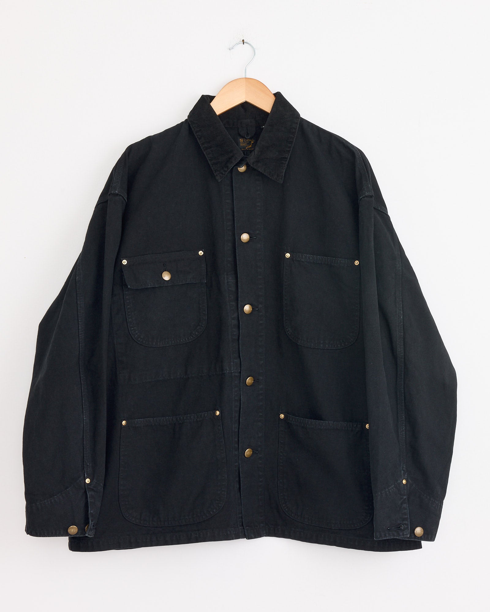 The orSlow Relax Fit Oxford Coverall Jacket in Black, featuring a collared button-up design and four front pockets inspired by classic workwear, is shown on a wooden hanger against a plain white background.
