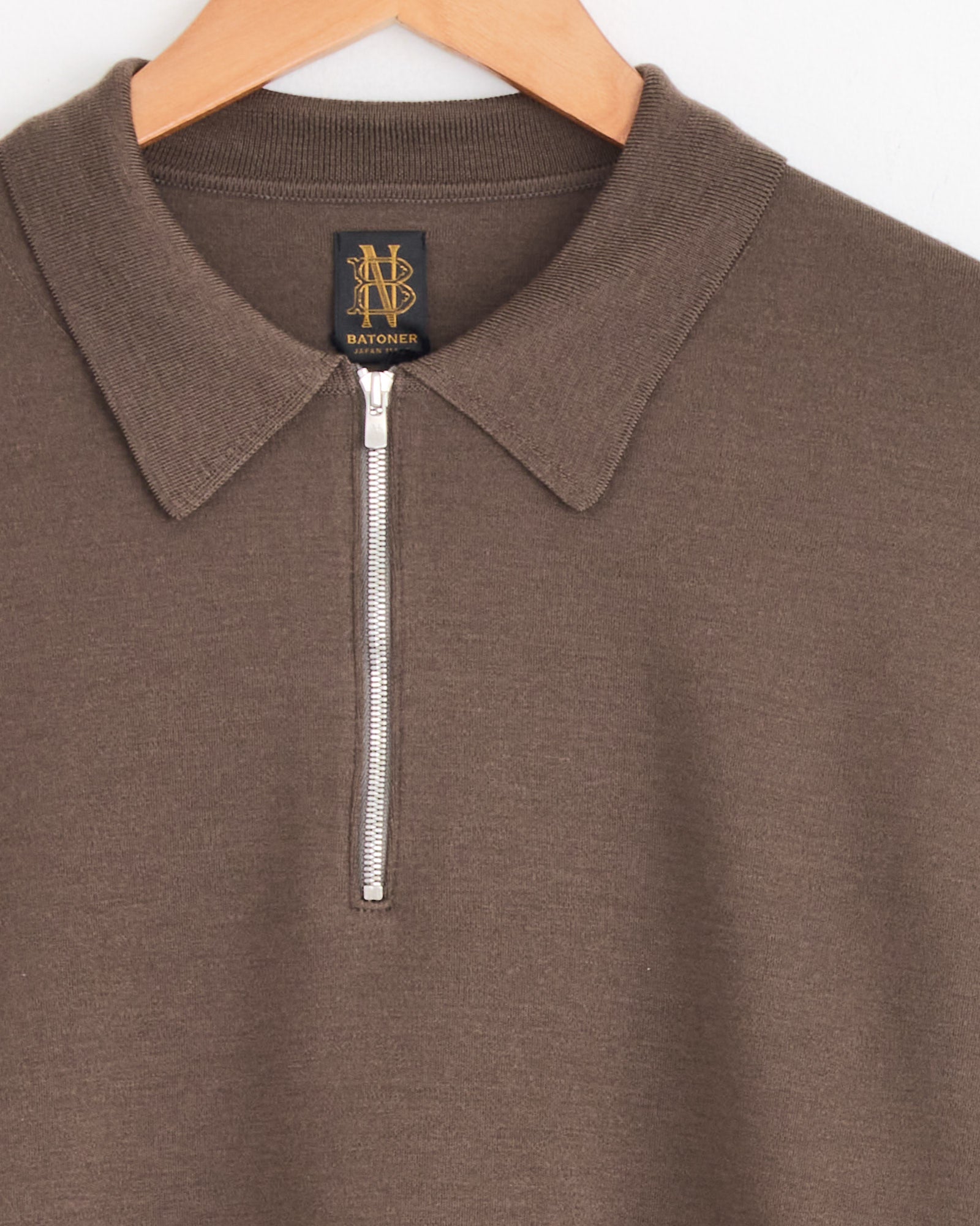 An olive Batoner Half Zip Polo hangs on a wooden hanger. Made in Japan, it features a silver half-zip closure and a black and gold brand label near the neckline.