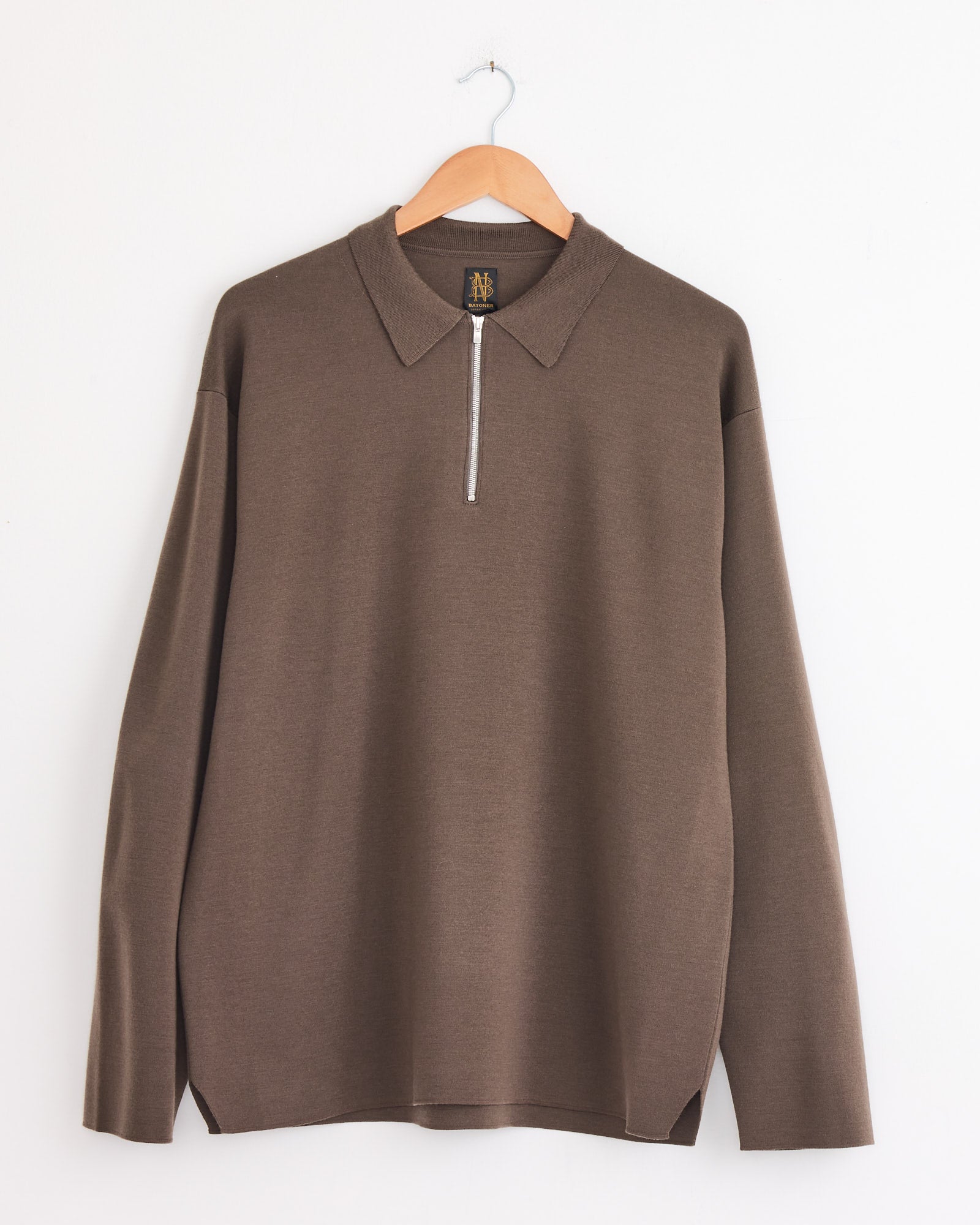 The Batoner Half Zip Polo in Olive, from the 32G Smooth Series and made in Japan, hangs on a wooden hanger against a white wall.