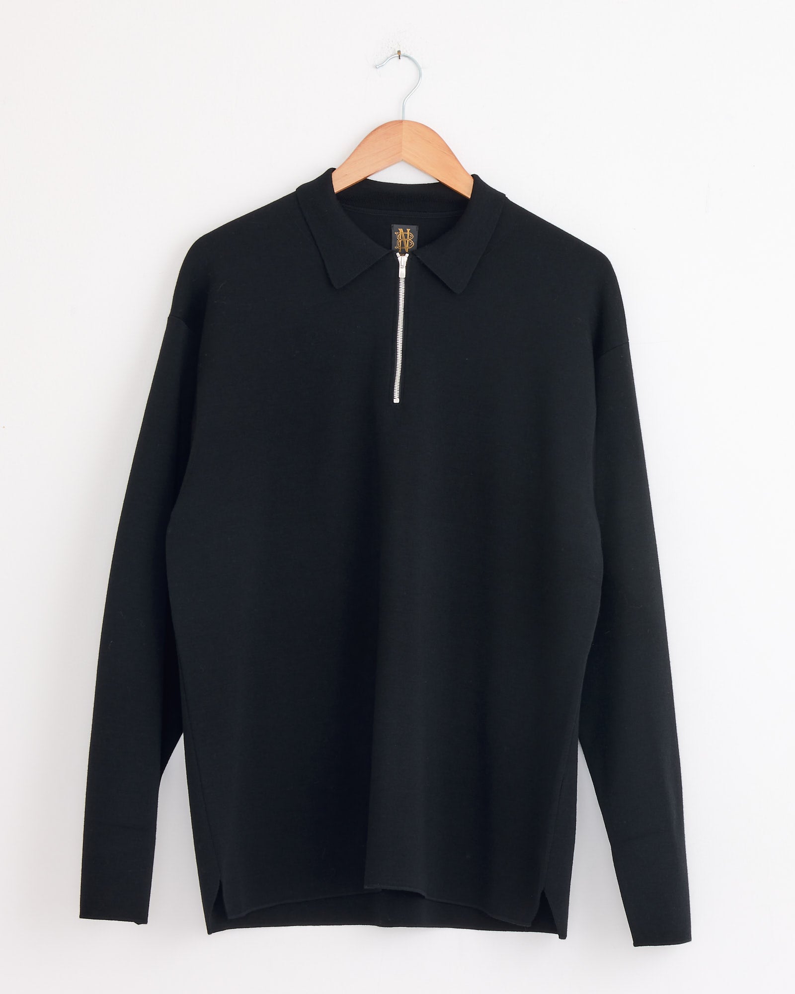 The Batoner Half Zip Polo in Black, a 100% wool long-sleeve shirt with a collar, hangs on a wooden hanger against a white wall.