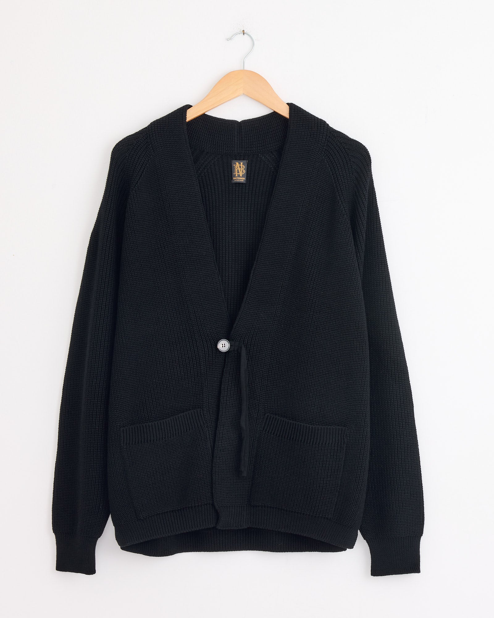 The Batoner Waso 1 Button Cardigan in Black, made in Japan from SOLIDWOOL yarn with long sleeves, two front pockets, and a single button closure, hangs on a wooden hanger against a plain white wall.
