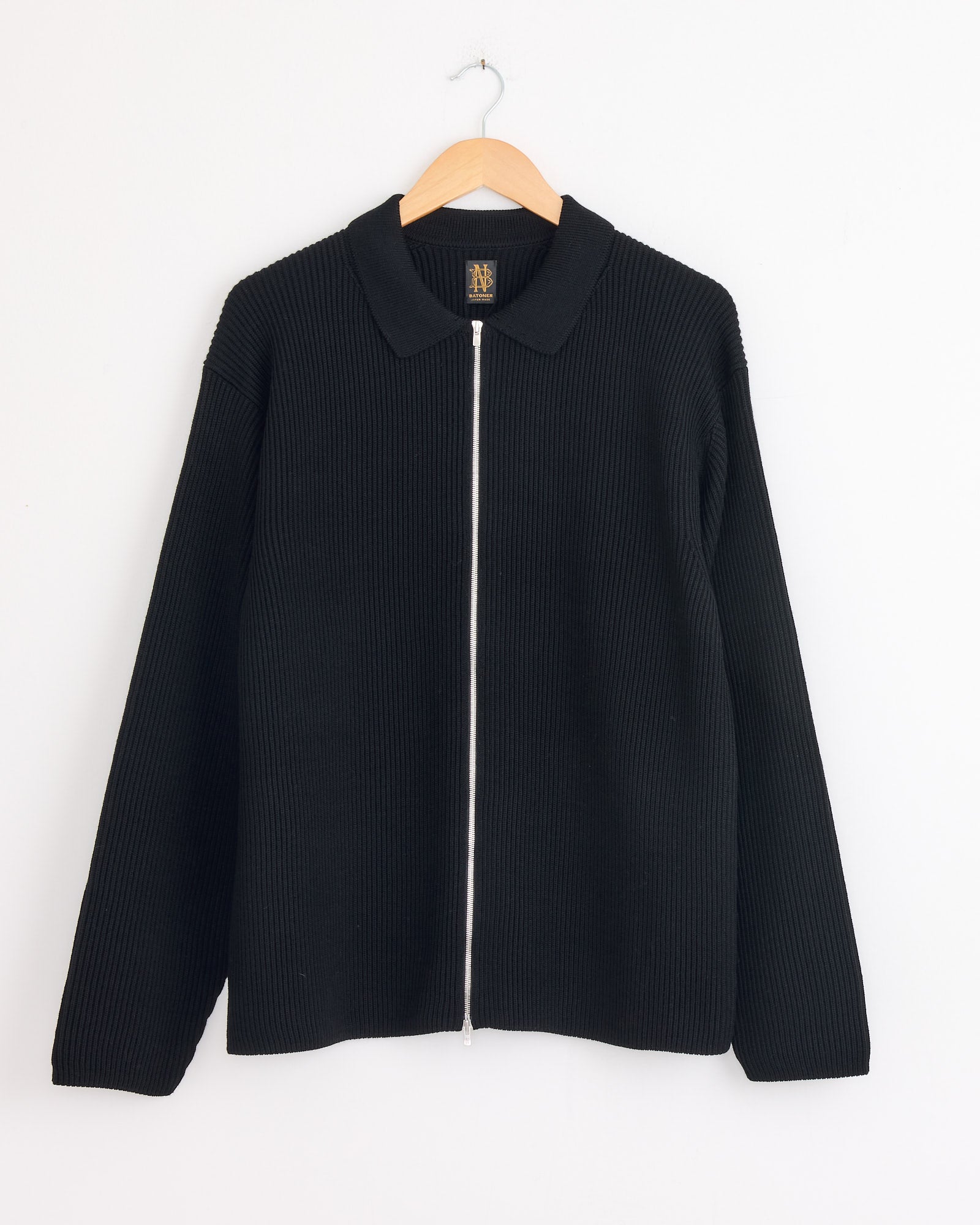 The Batoner Zip Up cardigan in Black, featuring a collar, long sleeves, and straight hem in premium SOLIDWOOL yarn, is shown on a wooden hanger against a plain white background.