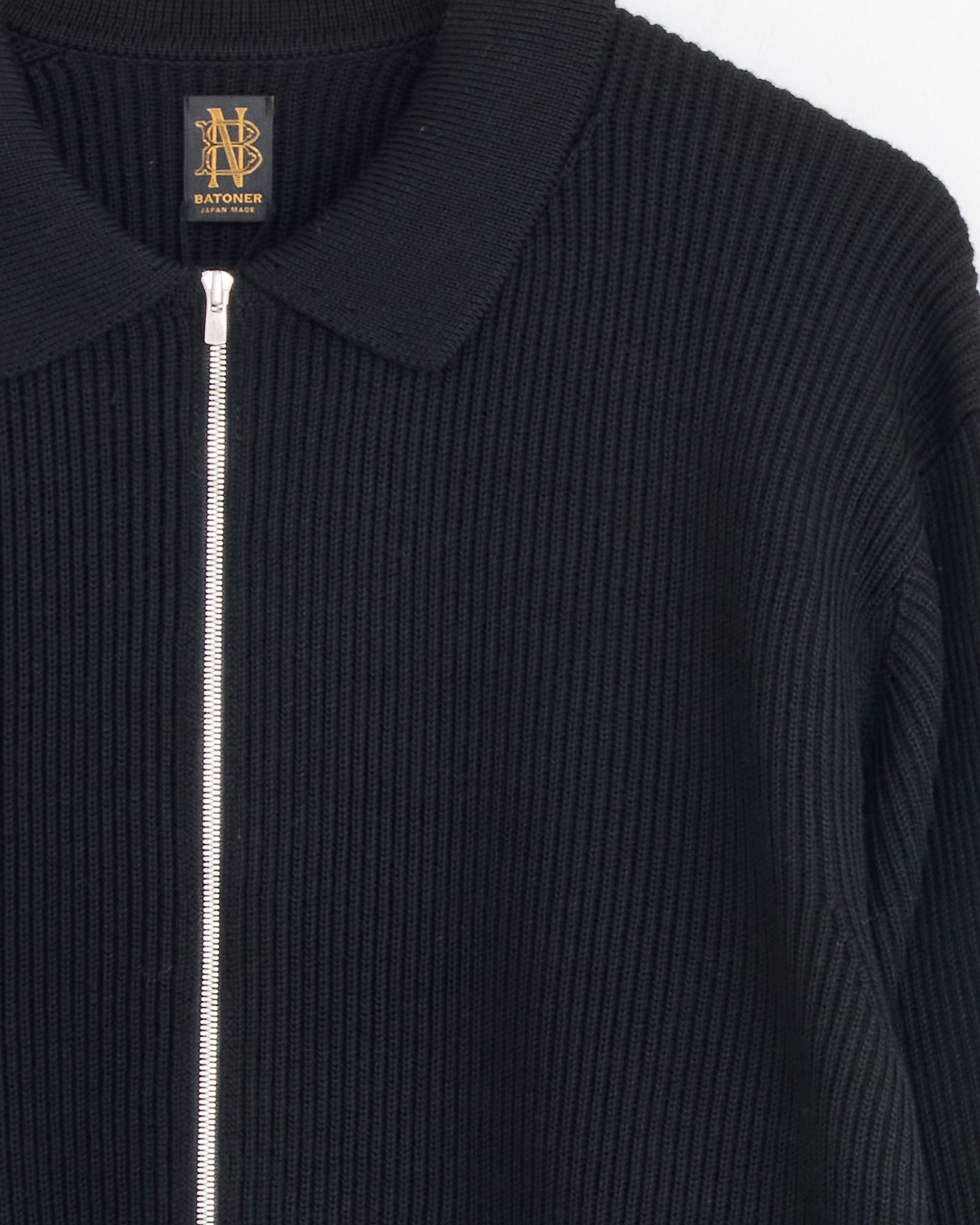Close-up of Batoner's Zip Up cardigan in Black, made in Japan, featuring a silver zipper, collar, and the brand’s gold and black label at the neckline.
