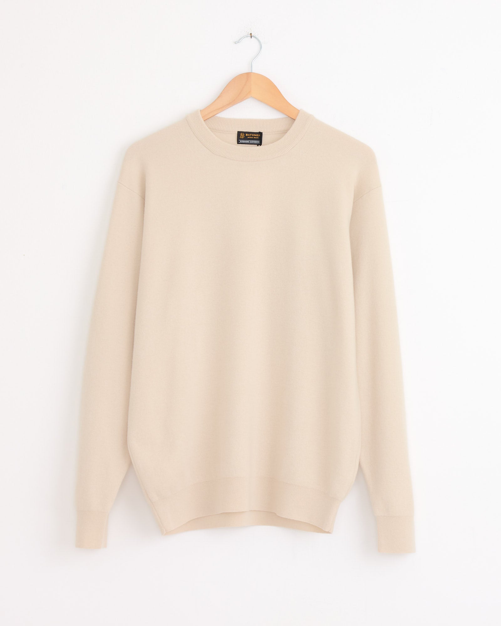 The Batoner Sunshine Crewneck in Natural, a luxurious light beige cashmere sweater, hangs on a wooden hanger against a white wall.
