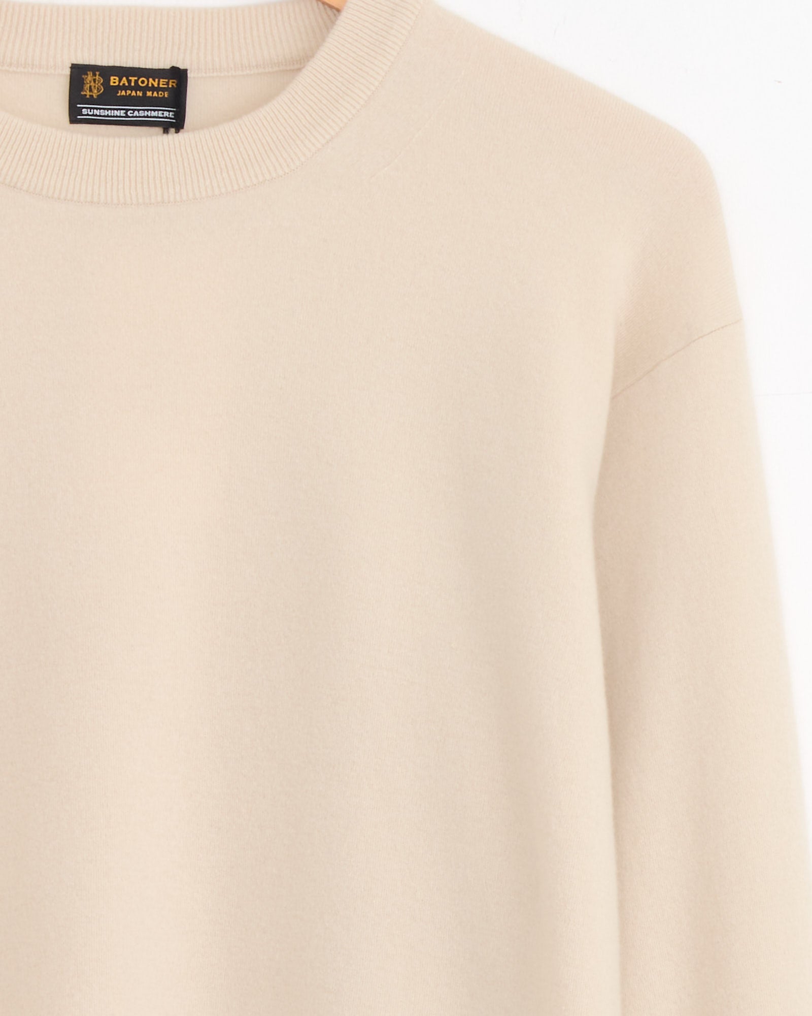 The Sunshine Crewneck in Natural by Batoner is a beige, long-sleeve cashmere sweater with a minimalist design and no visible patterns, featuring the label BATONER JAPAN MADE inside the collar.