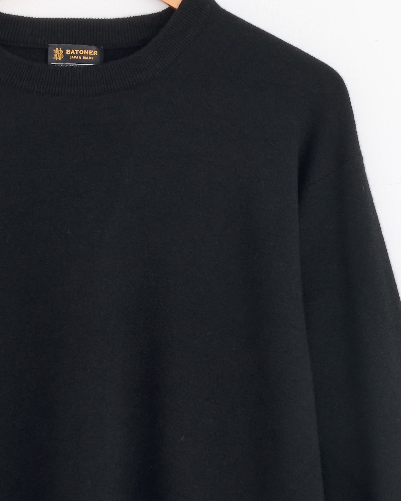 A close-up of the Batoner Sunshine Crewneck in Black hangs on a rack, highlighting the BATONER JAPAN MADE label and upper detail. This luxury sweater, crafted in Japan, is showcased against a plain white background.