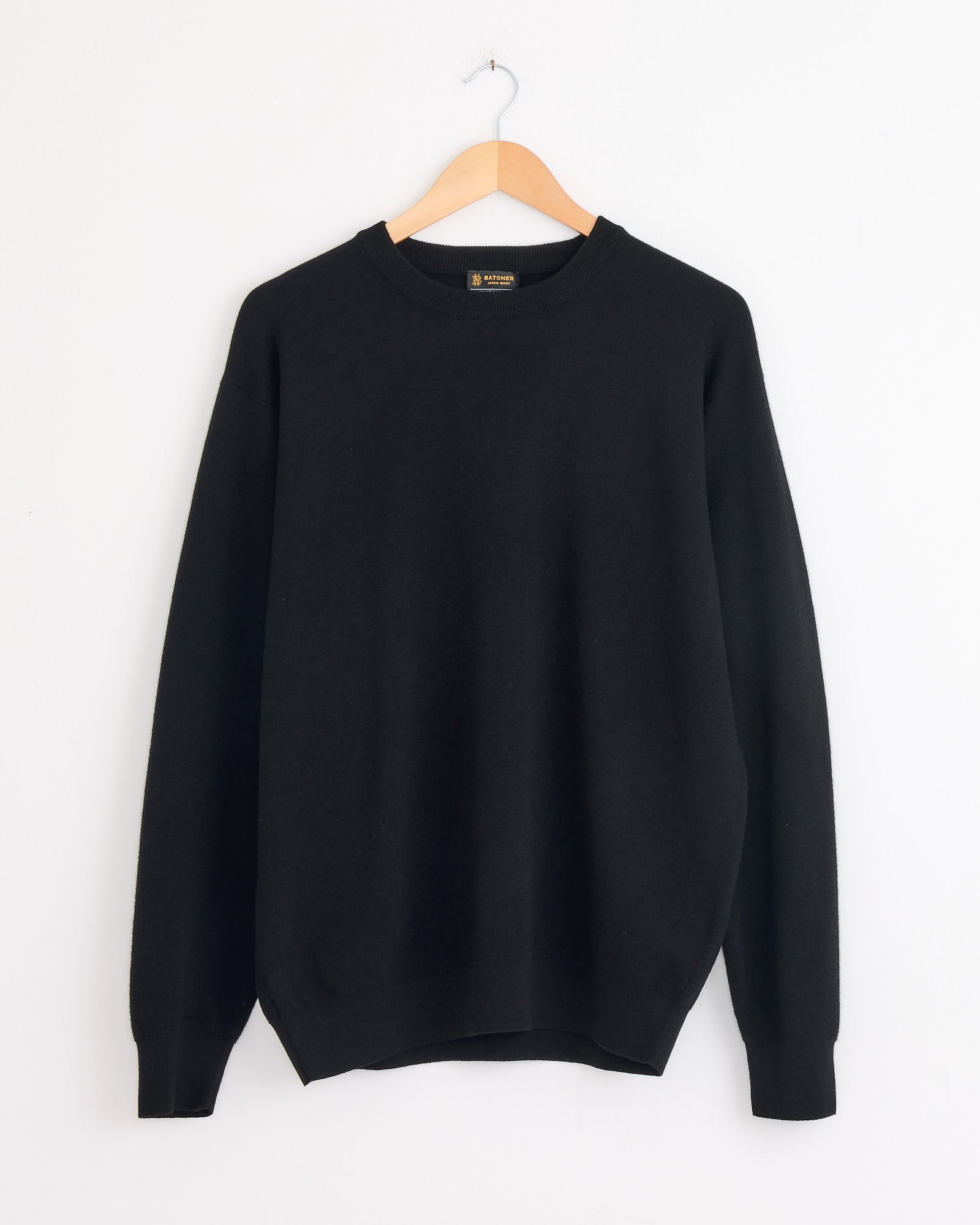 The Batoner Sunshine Crewneck in Black, a luxury cashmere sweater, is displayed on a wooden hanger against a white wall.