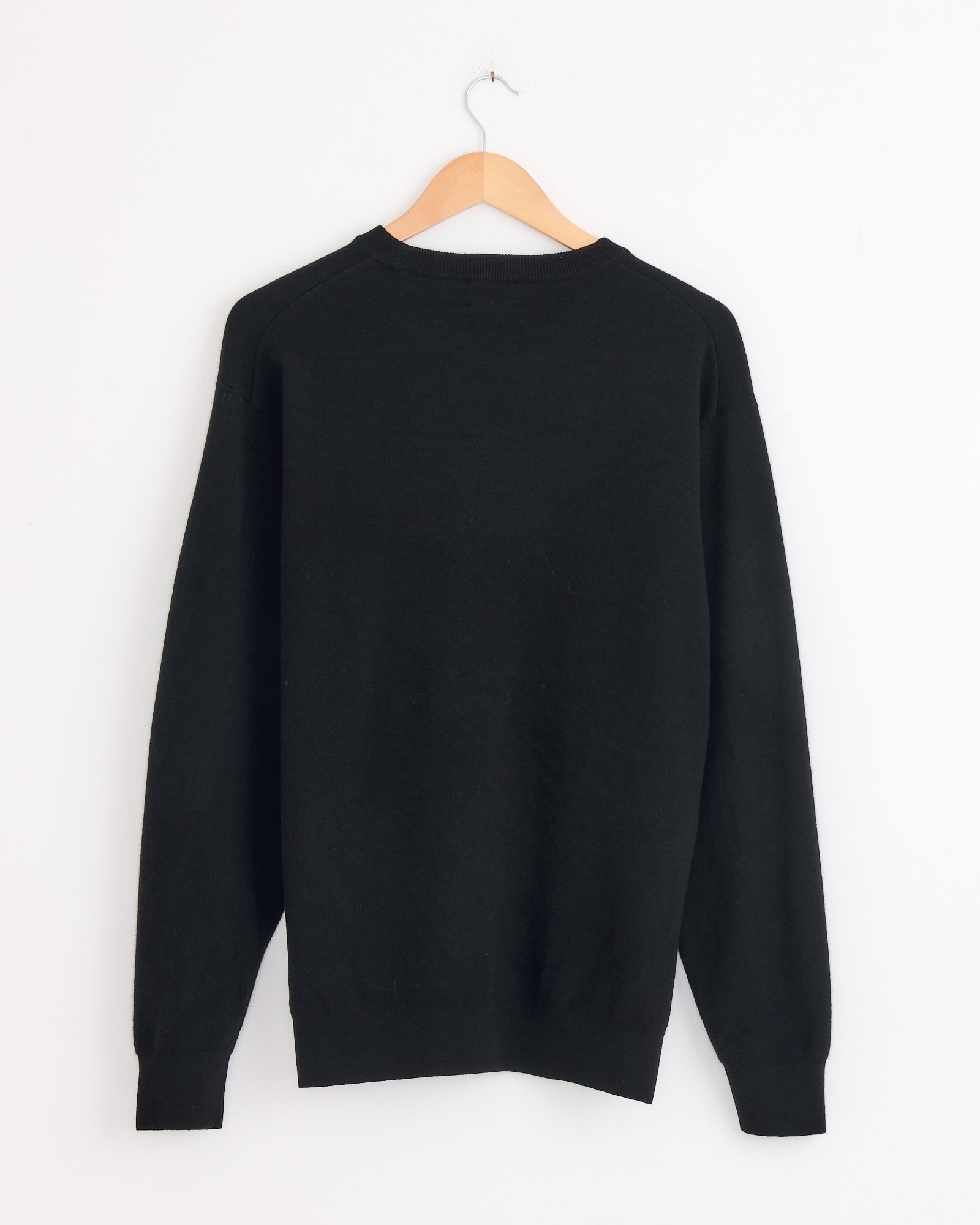 The Batoner Sunshine Crewneck in Black, a luxury cashmere sweater made in Japan, hangs on a wooden hanger against a white wall, displayed from the back.