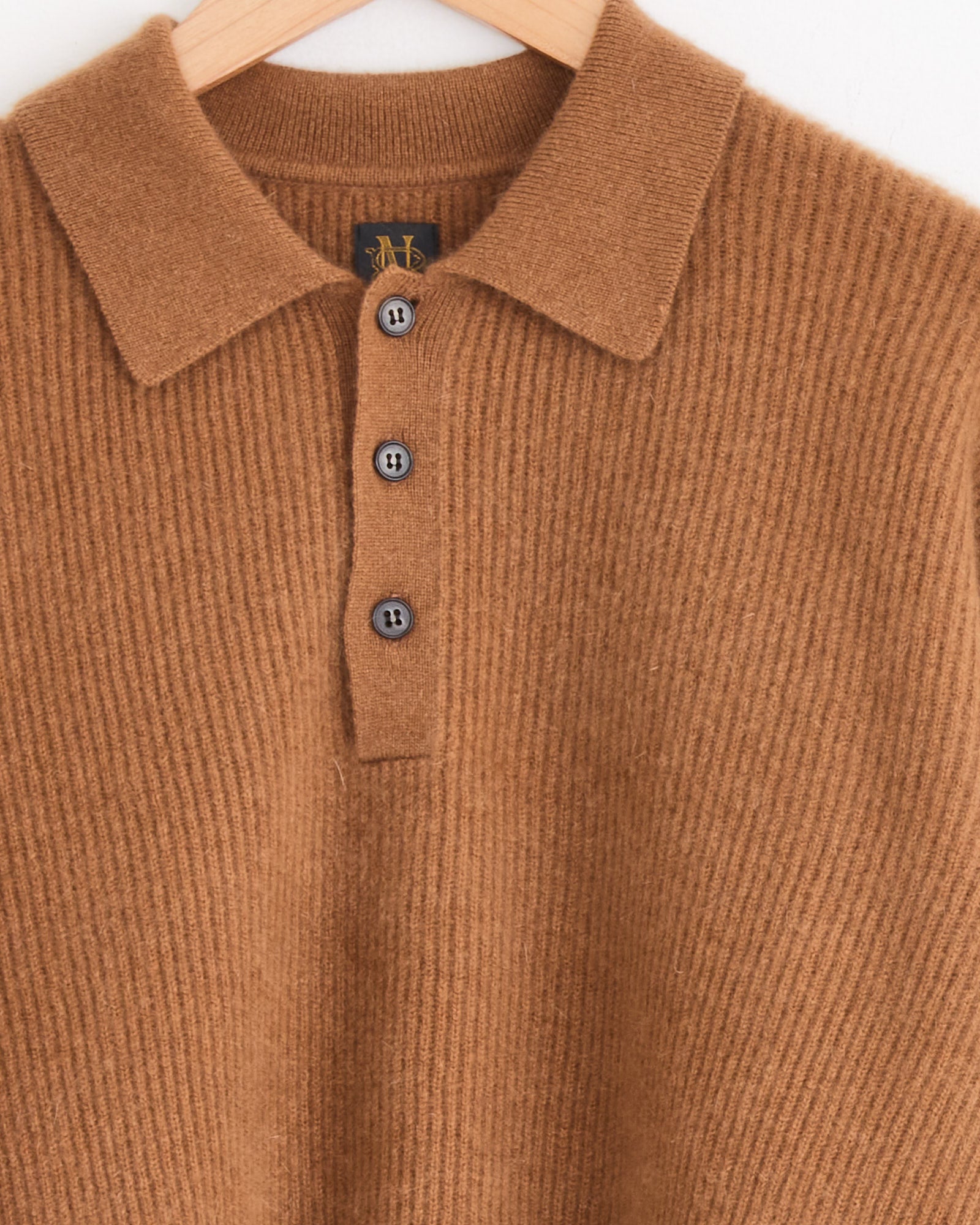 A close-up of the Batoner Fox Polo in Camel, a ribbed brown knit sweater with a collar and three dark buttons, hanging on a wooden hanger against a white background.