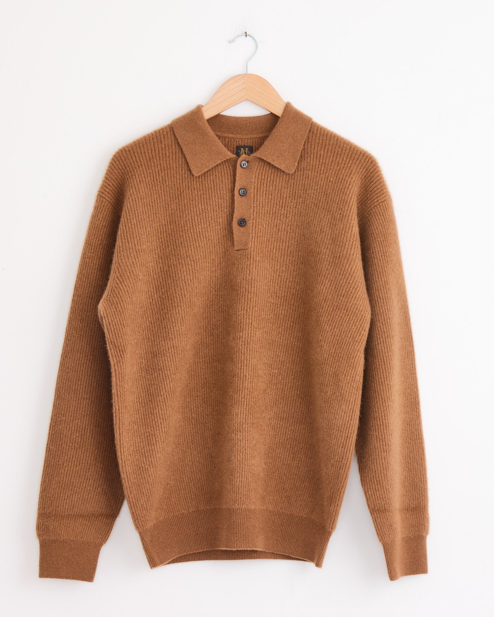 The Batoner Fox Polo in Camel is a long-sleeve knit sweater with a collar and three buttons, shown on a wooden hanger against a plain white wall.