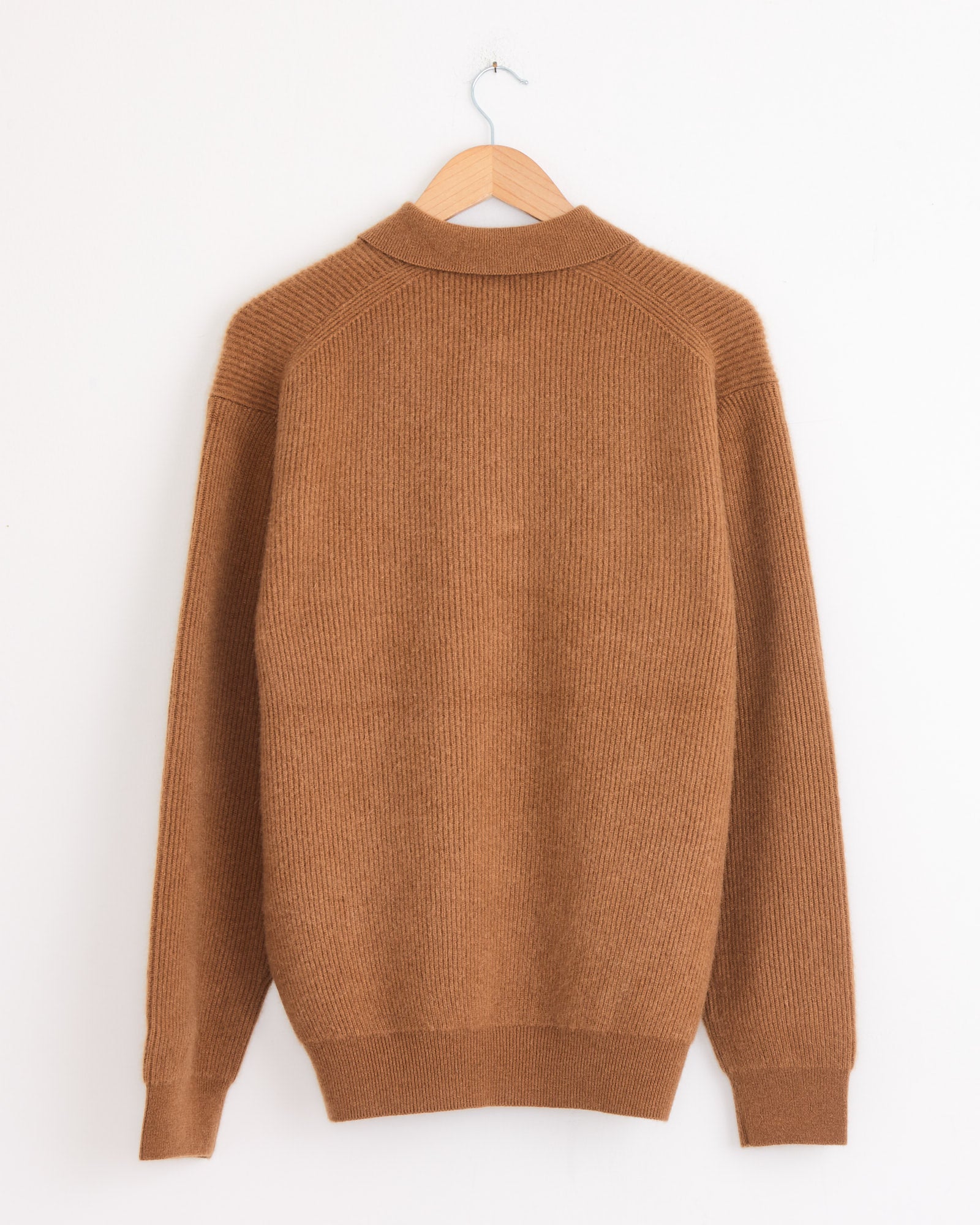 A Batoner Fox Polo in camel, featuring long sleeves and ribbed knit, hangs on a wooden hanger against a plain white wall, viewed from the back.