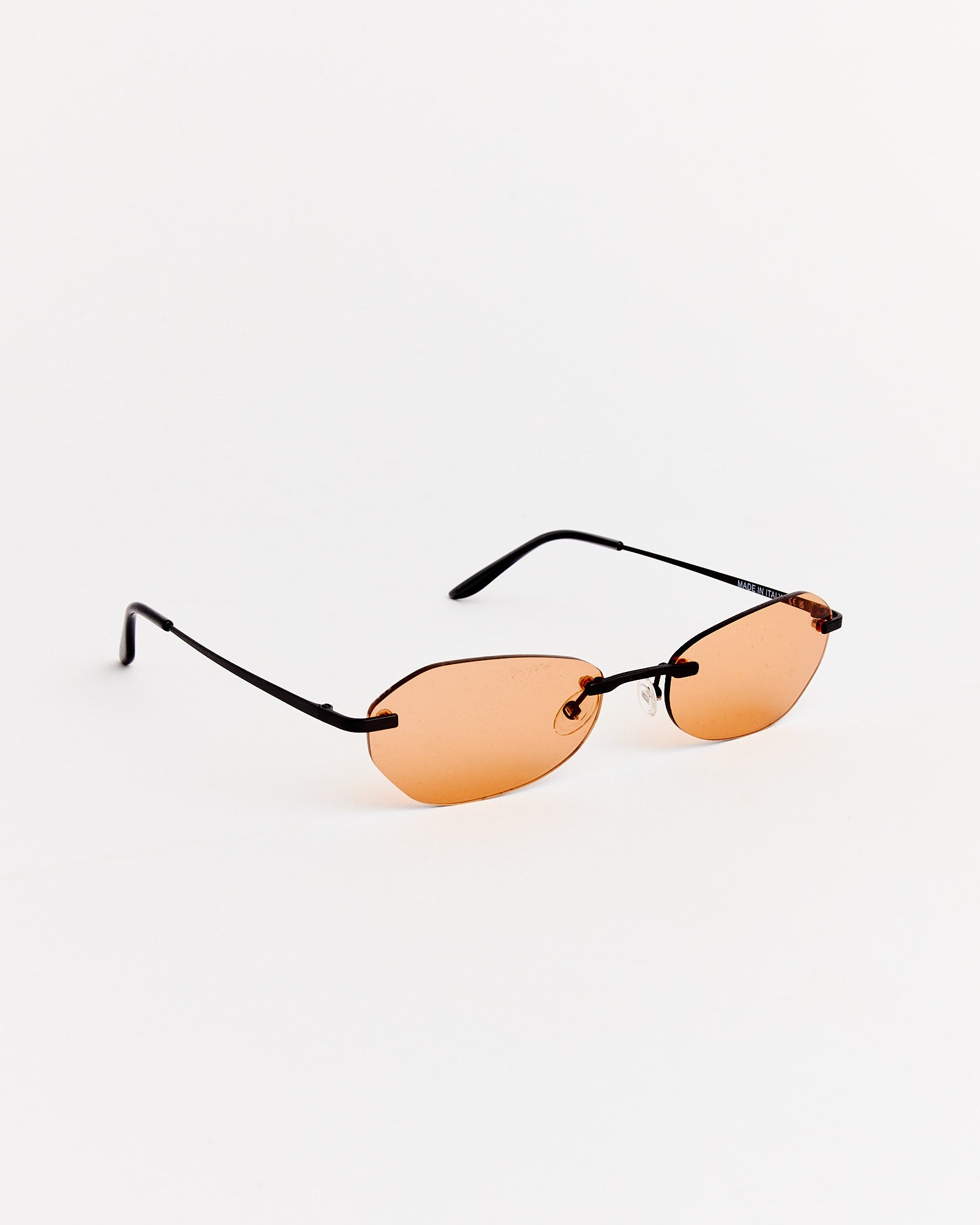 The Adorable Sunglasses in Graphite Spectra by Our Legacy feature rimless oval, orange-tinted lenses and thin black arms, shown against a plain white background.