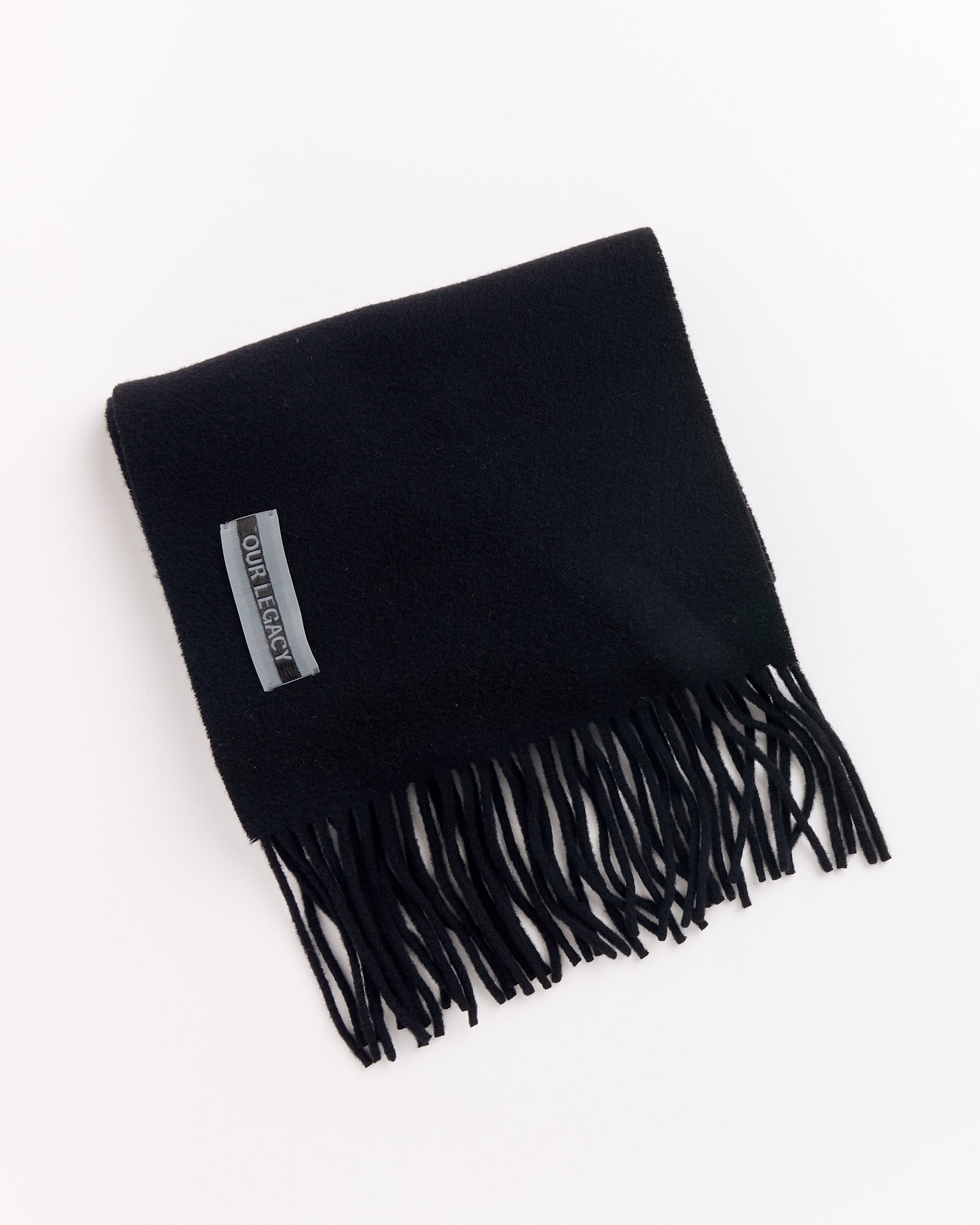The Estate Scarf in Black Double Wool by Our Legacy is neatly folded with fringe edges, featuring a sewn-on label reading "OUR LEGACY" in one corner. The scarf is displayed against a white background.